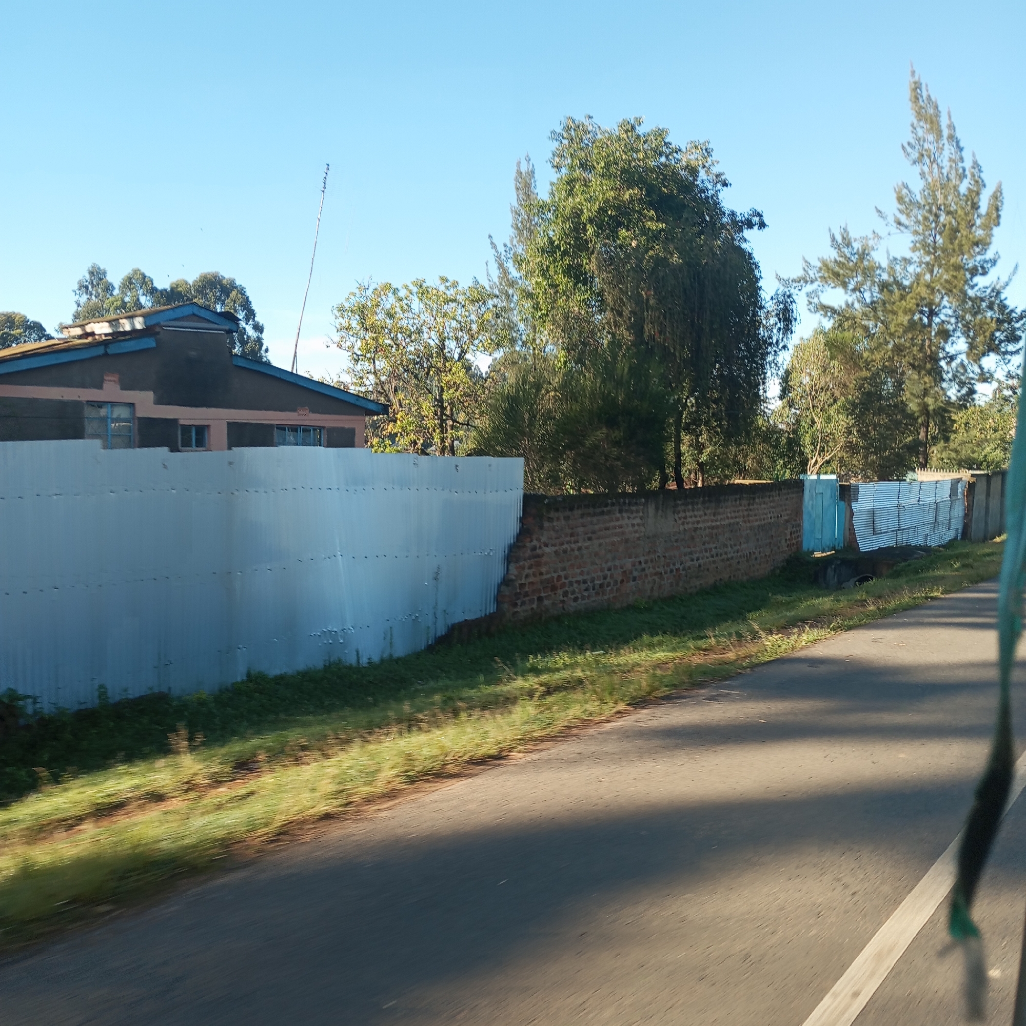 The image shows a street scene in Sheywe ward, Kenya. A two-lane asphalt road is visible, with a white dividing line. To the left of the road, there's a combination of fencing and walls. The fence closest to the viewer is made of corrugated metal sheets. This transitions into a brick wall. Further down, there's a blue wooden structure, then more corrugated metal fencing and a final brick column. Behind the fencing and walls is a single-story building with a dark roof, a light-colored exterior and blue trim. The sky is clear and blue. Trees are seen in the background. The photograph appears to have been taken from a moving vehicle.