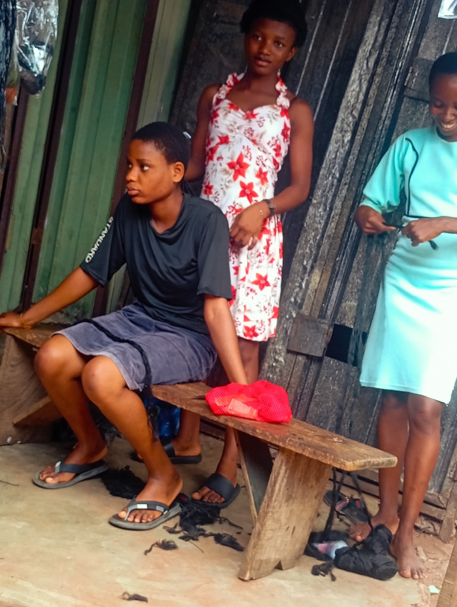 Three young Black females are standing and sitting on a wooden bench outside a building with green corrugated metal siding and a dark metal door. The female on the left is sitting on the bench, wearing a black t-shirt with white lettering on the sleeve and a gray skirt. She is wearing black flip-flops. A red mesh bag is on the bench next to her. The female in the middle is standing, wearing a white dress with red flowers. She is wearing black flip-flops. The female on the right is standing, wearing a light blue dress. She is barefoot. There are several pairs of shoes on the ground beneath the bench, and some hair clippings on the floor. The overall scene appears to be an outdoor market or street scene.