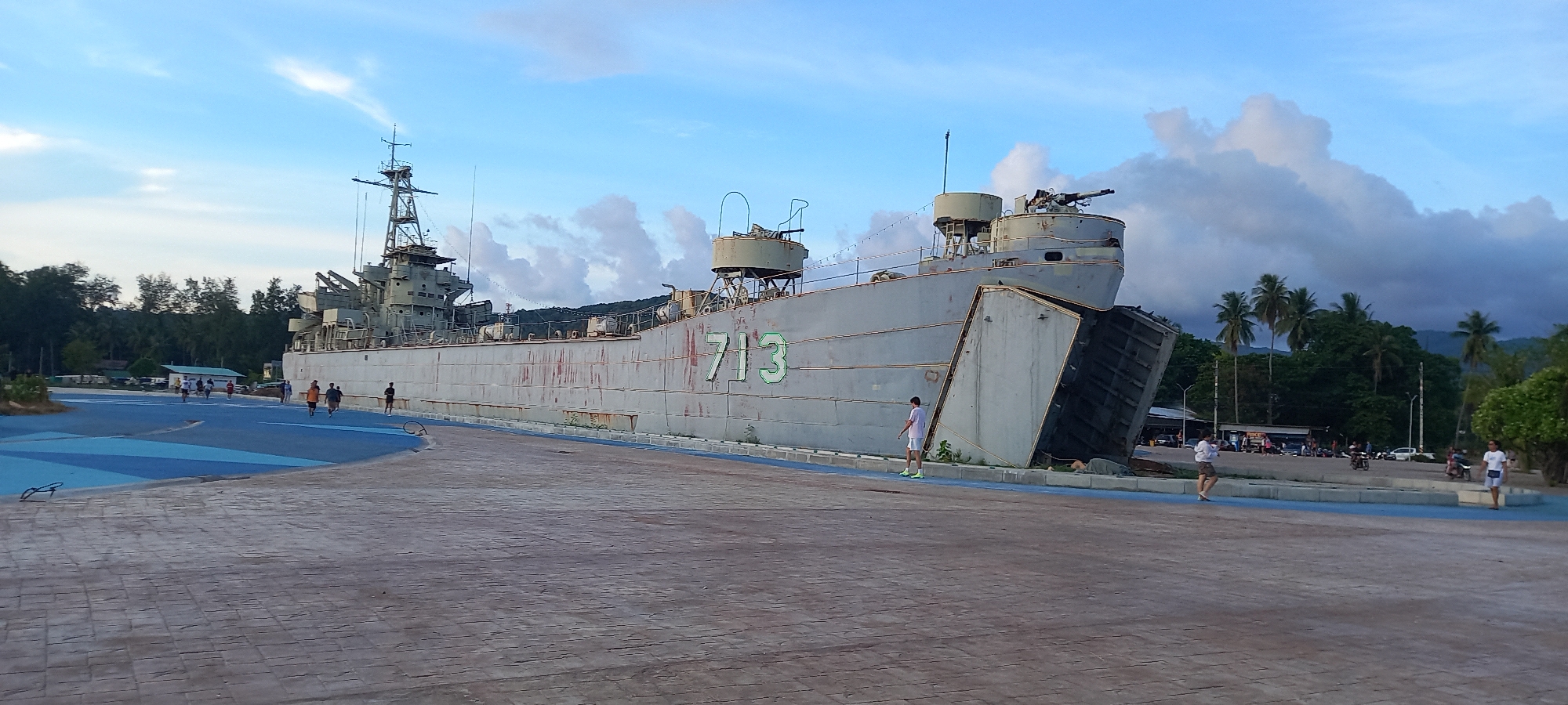 The image shows a large, gray, decommissioned naval vessel, bearing the number "713", permanently stationed on a paved area near Thong Sala in Thailand. The ship has a distinct landing craft design, with a bow ramp. Several people are walking around the vessel. The sky is partly cloudy. Trees and some buildings can be seen in the background. The area around the vessel appears to be a park or public space with blue painted pathways.