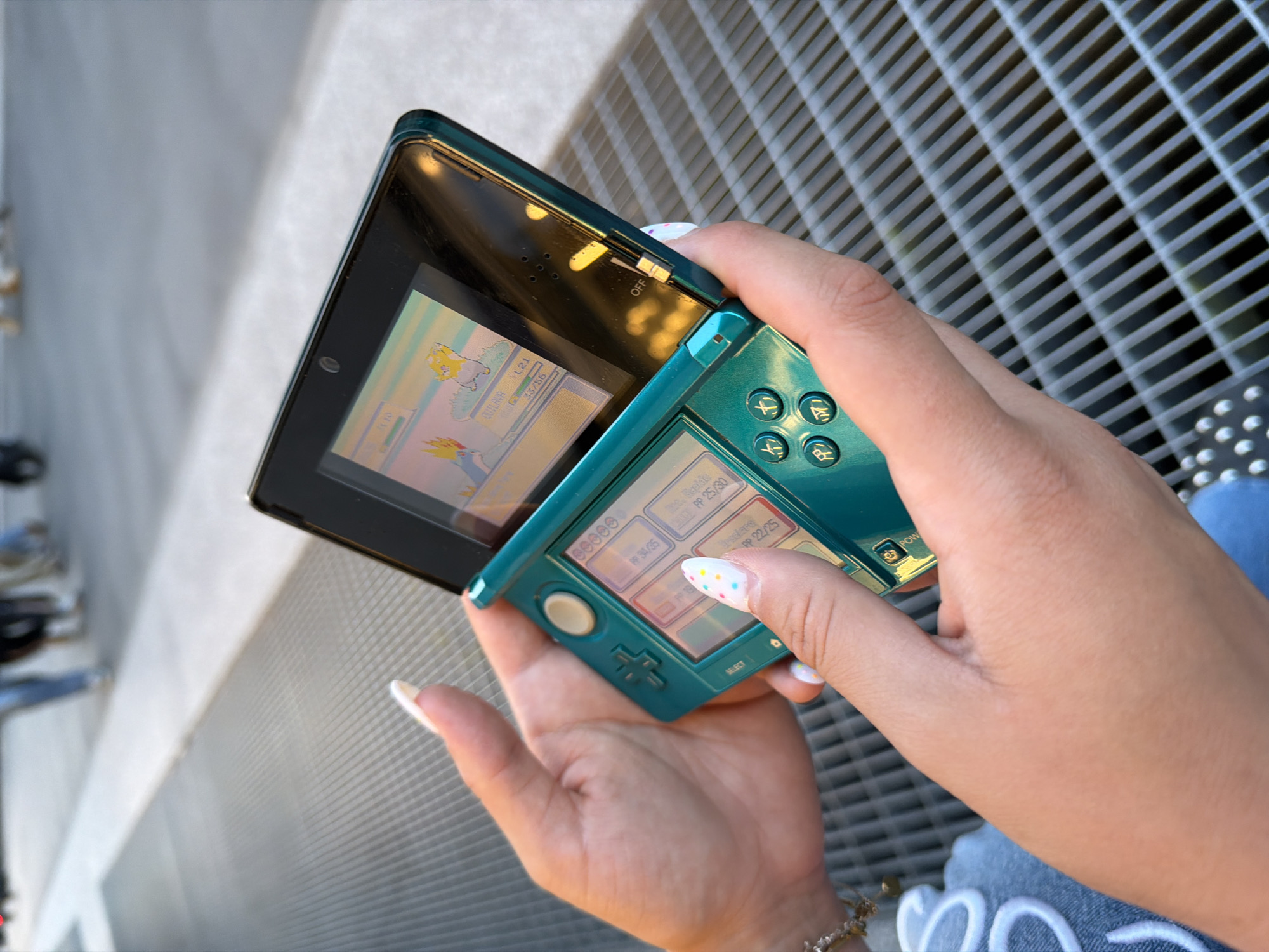 A close-up, eye-level shot shows a person's hands holding an open, teal-colored Nintendo 3DS. The top screen displays a pixelated game scene with two colorful, cartoonish creatures. The bottom screen shows game text and menu options. The person's fingernails are painted with a white base and multi-colored dots, and a silver bracelet is visible on the left wrist. The 3DS rests on a metal grate. In the blurred background, several people walk on a sidewalk with a white line marking a path. The scene appears to be outdoors, likely in an urban environment.