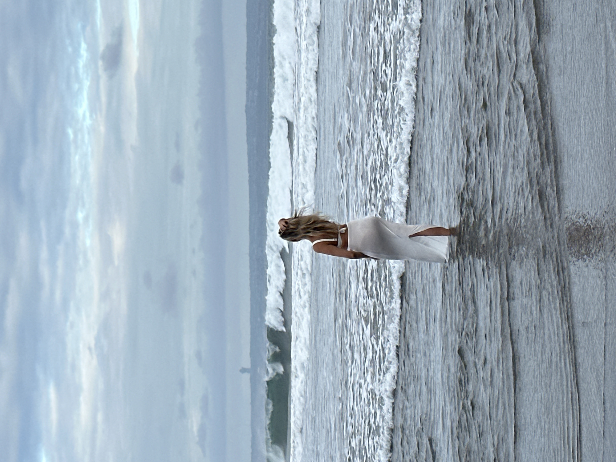 A person wearing a white dress stands in the ocean at a beach in North Kuta, Indonesia. The person is facing away from the camera, looking out at the waves. The sky is cloudy and the water is choppy. There is a structure visible in the distance on the horizon. The image appears to be taken during the daytime.
