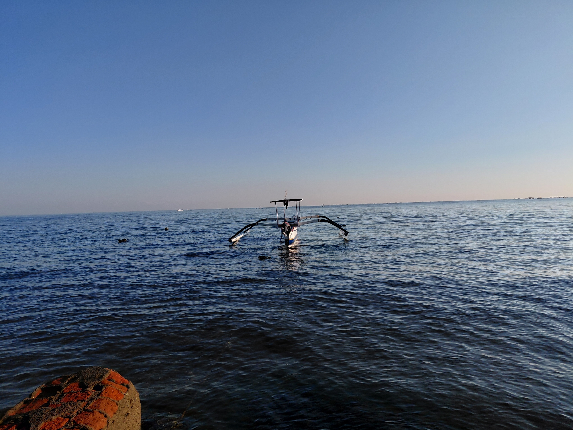 The image depicts a serene seascape at sunrise or sunset in Buleleng, Indonesia. The calm, deep blue ocean stretches towards a clear, pale blue sky, with a subtle gradient suggesting the low position of the sun. In the mid-ground, a traditional Indonesian outrigger boat, known as a "prahu," is the central subject. The boat has a white hull with black outriggers extending outwards, and a small canopy structure at its center. The boat appears to be stationary on the water. In the foreground, a partially submerged, rough-textured object, likely a weathered piece of concrete or rock with visible reddish-orange brick fragments, anchors the lower-left corner of the frame. The water surface is gently rippled, reflecting the soft light. Two dark, small shapes, possibly birds or buoys, are visible in the water to the left of the boat. The horizon line is sharp and well-defined, with faint outlines of distant landmasses or other boats visible on the far right. The overall impression is one of tranquility and natural beauty.