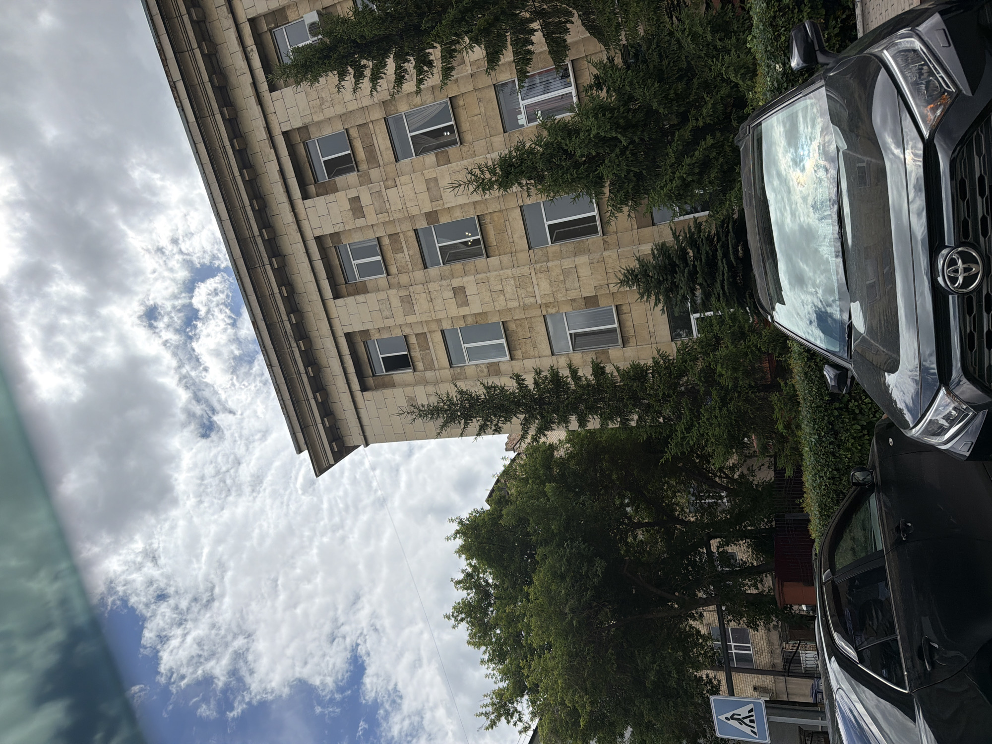 The image captures an outdoor scene featuring a building with a stone facade and numerous windows, partially obscured by lush green trees. In the foreground, a dark-colored car is visible, with its reflection showing the sky and clouds. The sky is partly cloudy, with a mix of white and gray clouds against a blue background, suggesting daytime.
The building appears to be a multi-story structure, possibly an administrative or public building, given the location context provided as Qarağandı city administration, Kazakhstan. The architecture is characterized by its stone blocks and regular window placements. The trees, likely conifers and deciduous varieties, add a natural element to the urban setting, with their branches reaching towards the building and the sky.
The car in the foreground is a modern vehicle, seen from a side angle, highlighting its reflective surfaces. The reflection on the car's windshield shows a bright, cloudy sky, indicating it's likely midday or afternoon. The overall impression is one of a calm, everyday scene in a city environment. There are no people visible in the frame.
A small, blue and white traffic sign is visible on the right side of the image, indicating a pedestrian crossing. This further reinforces the urban setting. The image appears to be taken from a low angle, possibly from another vehicle or from the ground, looking upwards towards the building and the sky. The lighting suggests it's a bright day, despite the presence of clouds.