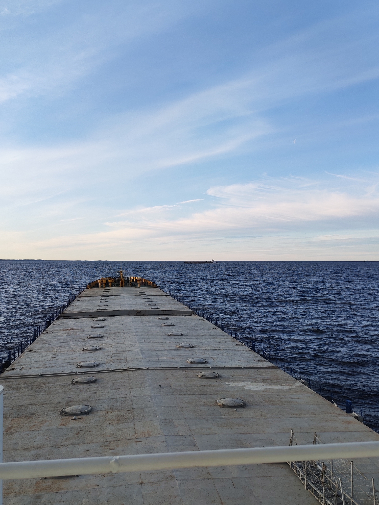View from a barge or large boat with a concrete deck, showing a body of water and horizon under a blue sky with wispy clouds and a crescent moon. The deck of the boat has several circular manhole covers. There are other ships in the distance. The image appears to be taken at or near dusk. This scene is not in Novaya Svobodka, Russia. There are no people visible in the image.