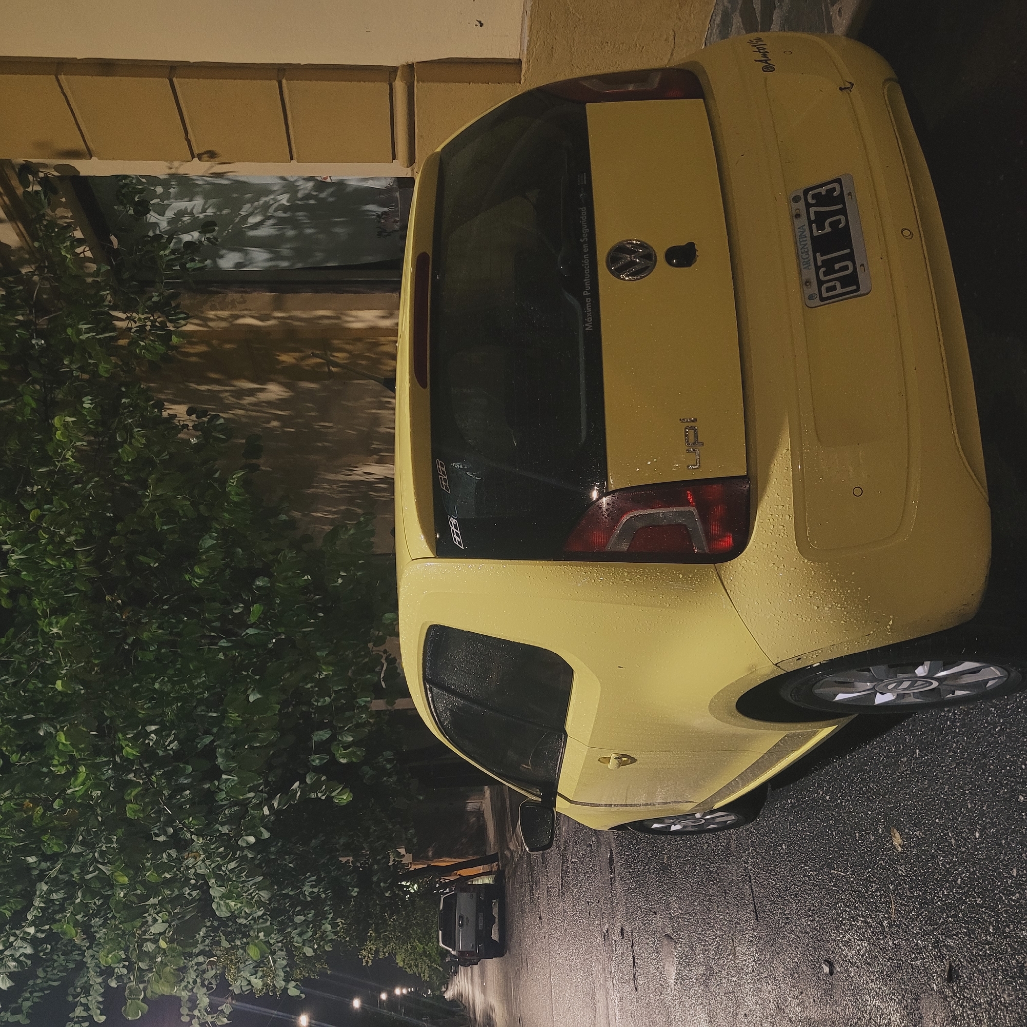 A yellow Volkswagen Up! car, bearing license plate PGT 573 from Argentina, is parked on a wet, asphalt street in San Fernando del Valle de Catamarca. The car is oriented facing away from the camera. A building facade is visible next to the car with a window reflecting light. A tree with dense foliage is positioned adjacent to the building. In the distance, other cars are parked along the street, illuminated by streetlights.