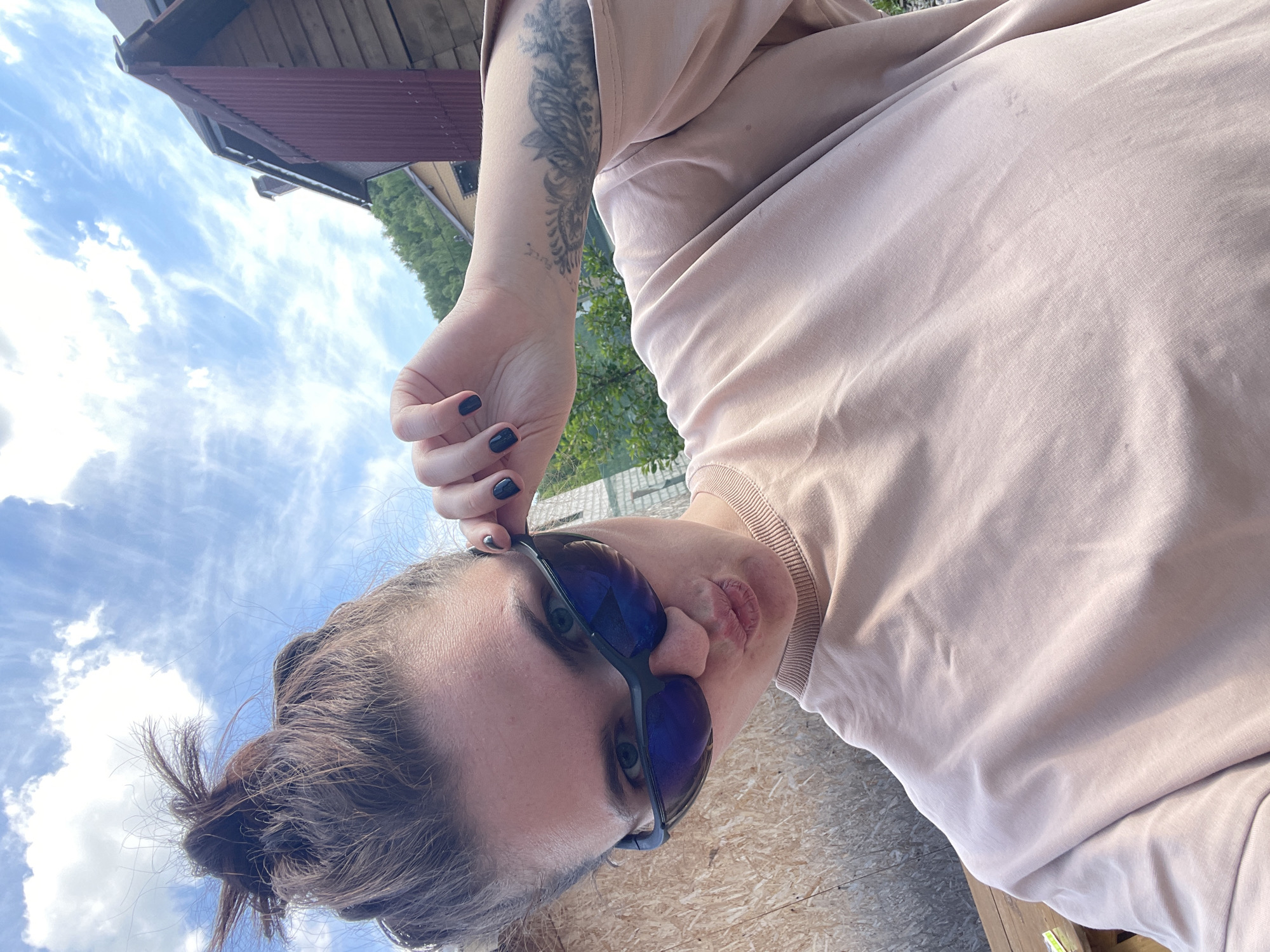 The image shows a person wearing sunglasses and a light pink or beige outfit, with a tattoo visible on their arm. The person is outdoors, with a view of a partly cloudy sky and a building or structure in the background that appears to be in Мосметаллоконструкция Онт, Russia. The person's hand is raised near their sunglasses. The ground or surface beneath them appears to be wooden.