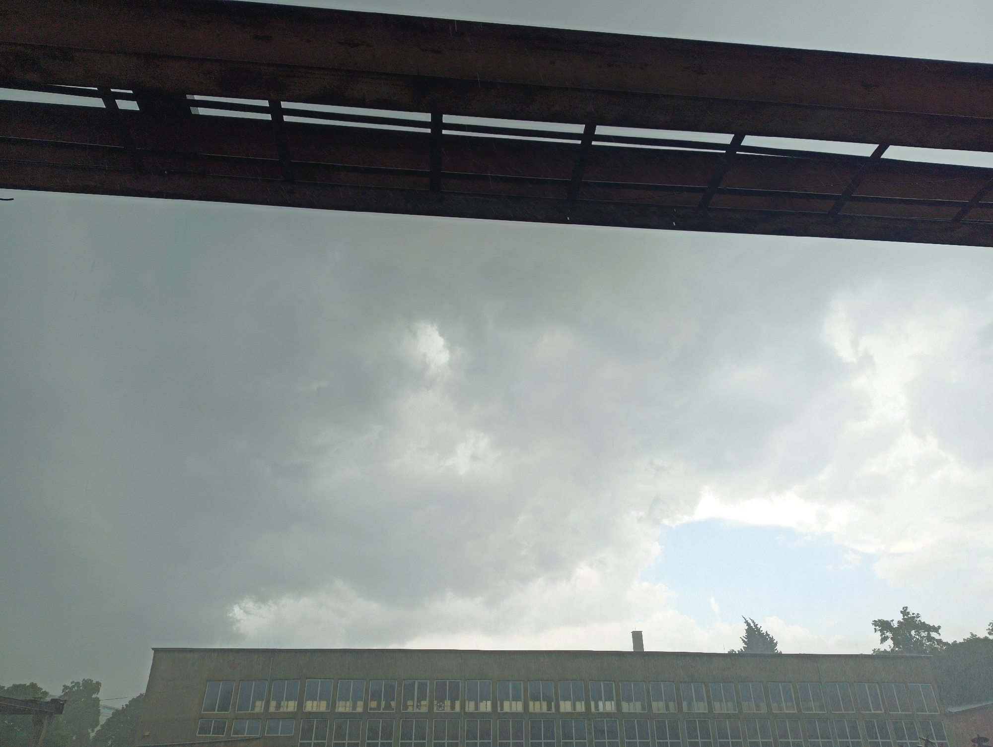 The image captures a view of a grey, overcast sky above a low, rectangular building with many windows. The building appears to be old and utilitarian, with a chimney on its roof. Trees are visible to the right of the building. A dark, possibly rusty, metal structure is in the foreground, obscuring the top of the view. Raindrops are visible on the metal structure. The location is in the city of Unknown, Unknown.