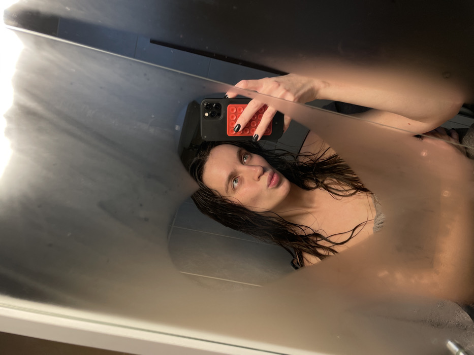 A mirror selfie reveals a young woman with wet, dark hair and fair skin, pouting slightly at the camera. She's wearing a thin-strapped top or possibly a towel. Her nails are painted black. She's holding a black smartphone with a red, textured grip on the back. The mirror's surface is partially steamed up or foggy, creating a blurred effect, especially around the edges. The bathroom setting is simple, with gray tiles visible in the background.
