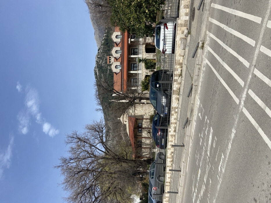 The image displays a daytime exterior view of the Monastery Podmaine complex in Budva, Montenegro, observed from the Jadranska magistrala (E80/E65) road. The scene is illuminated under a clear blue sky with a few scattered white clouds.
In the foreground, an asphalt road is visible with faded white pedestrian crossing markings and other partial road lines. A curb separates the road from a narrow strip containing five dark grey bollards; two are upright, and three are bent forward and resting on the ground.
Behind the strip, a low stone wall topped with a dark metal fence encloses a parking area. Four vehicles are parked parallel to this fence: a partially visible dark grey Skoda Octavia sedan on the far left, a dark blue sedan, a dark grey minivan, and a partially visible white sedan on the far right.
Behind the parked vehicles, two prominent stone buildings with red-tiled roofs constitute the monastery complex. To the left, a smaller stone building features a central dome topped by a cross, identifying it as the monastery church. To its right, a larger two-story stone building presents a red-tiled roof with three distinct arched dormer windows and a ground-level balcony with stone columns and railings.
The mid-ground includes several large deciduous trees, mostly bare of leaves, and some evergreen foliage near the buildings. The background is dominated by rocky, sparsely vegetated mountains rising beneath the clear sky. No individuals are present in the scene.