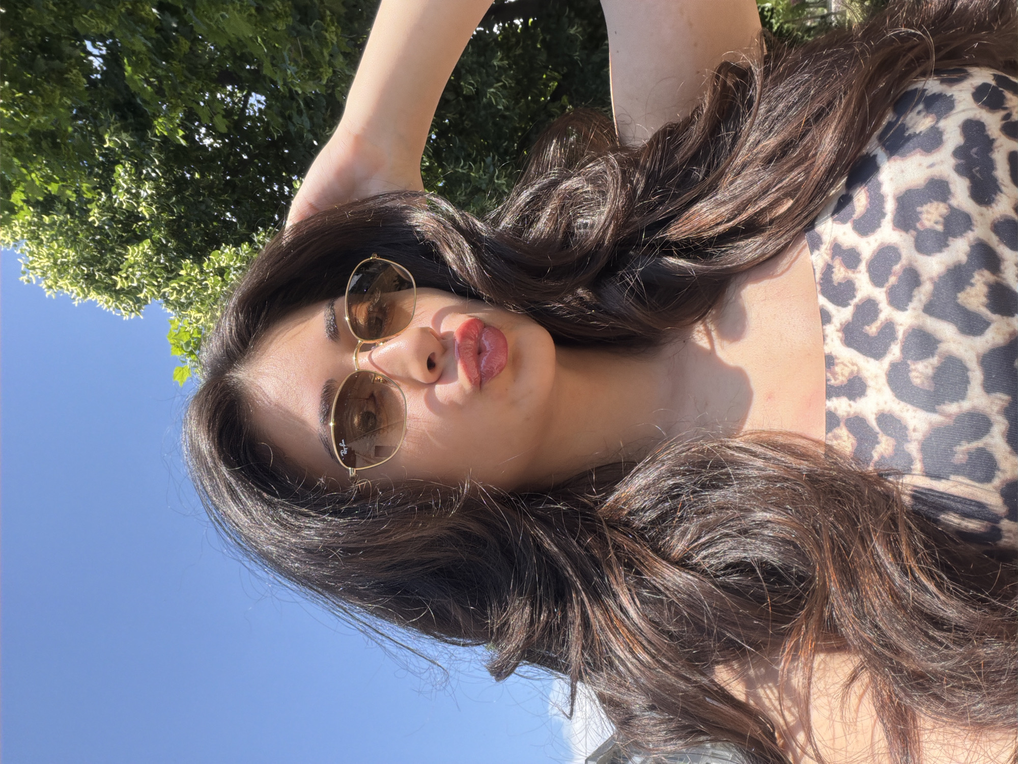 A woman with long, brown hair and sunglasses poses against a backdrop of blue sky and green foliage. She is wearing a leopard-print top. The location in Blagoevgrad, Bulgaria is not discernible from the image.