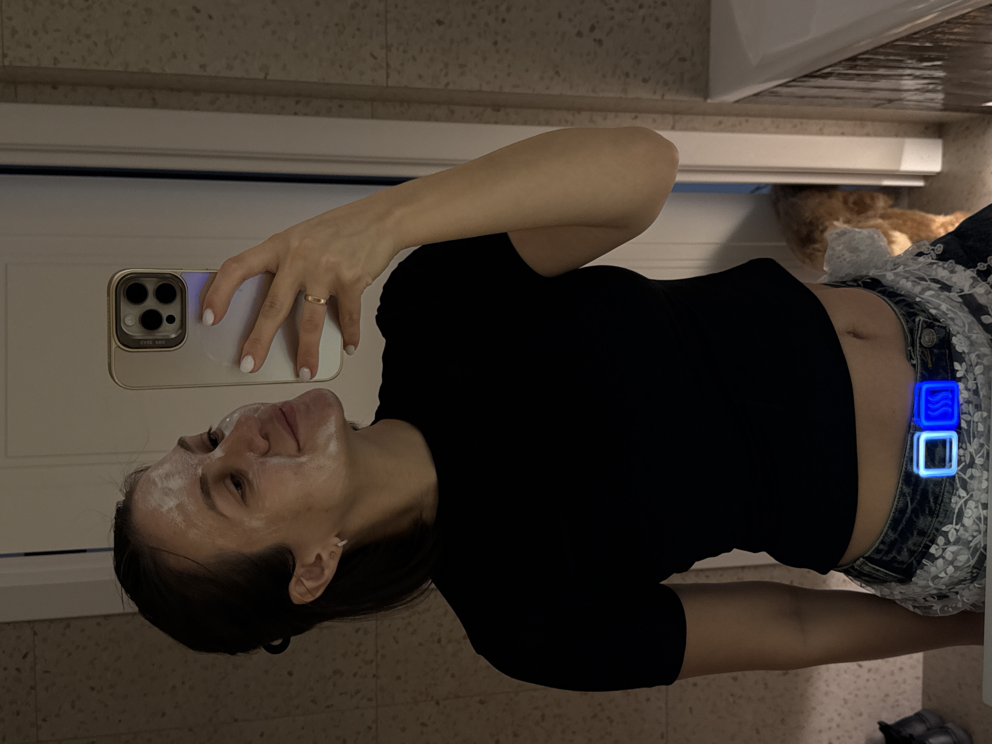 A young woman in Pervomaysky, Russia, captures a mirror selfie indoors, likely in a bathroom setting. The image, initially rotated 90 degrees, shows her standing upright, holding a light-colored smartphone horizontally in her right hand. Her hand features white nail polish and a gold band on her ring finger. The phone's reflection shows reversed text that reads "OYSE BBO" or similar, visible on its case.
She is wearing a black short-sleeved top that exposes her midriff and navel, paired with blue denim shorts or jeans adorned with a white, lacy pattern. On her right hip, two distinct glowing blue square devices are clipped to her waistband; one displays a wavy line icon, while the other is an empty square outline.
The woman's face is partially covered with a white substance, characteristic of a face mask or cleanser, applied to her forehead, cheeks, and chin. She gazes upwards towards the phone, her expression a gentle, relaxed smile, suggesting she is in the midst of a skincare routine. The background reveals light-colored tiled walls or flooring and a white fixture, possibly a sink or cabinet. A blurred, reddish-brown object, indistinct but possibly a pet or furniture, is visible further in the background. The subdued indoor lighting suggests the event is taking place in the evening.