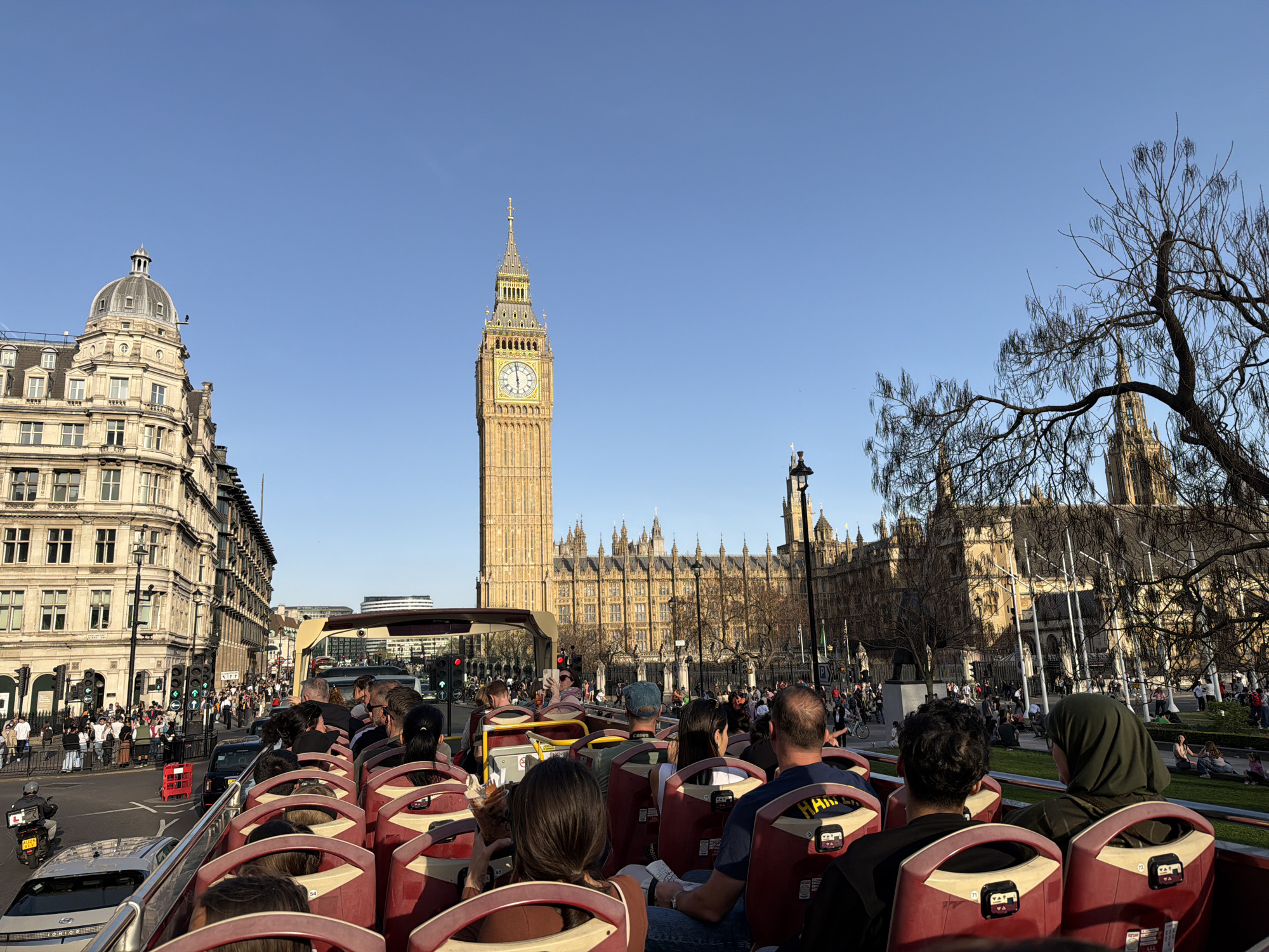 The image shows a view from the upper deck of a red open-top tour bus in London, United Kingdom. The bus is filled with passengers, with the backs of their heads and shoulders visible. In the background, the Houses of Parliament and Elizabeth Tower (Big Ben) are prominent. A building with a dome is on the left. A bridge is visible over the road. People are walking along the sidewalks. Trees are bare or budding. The sky is clear and blue.