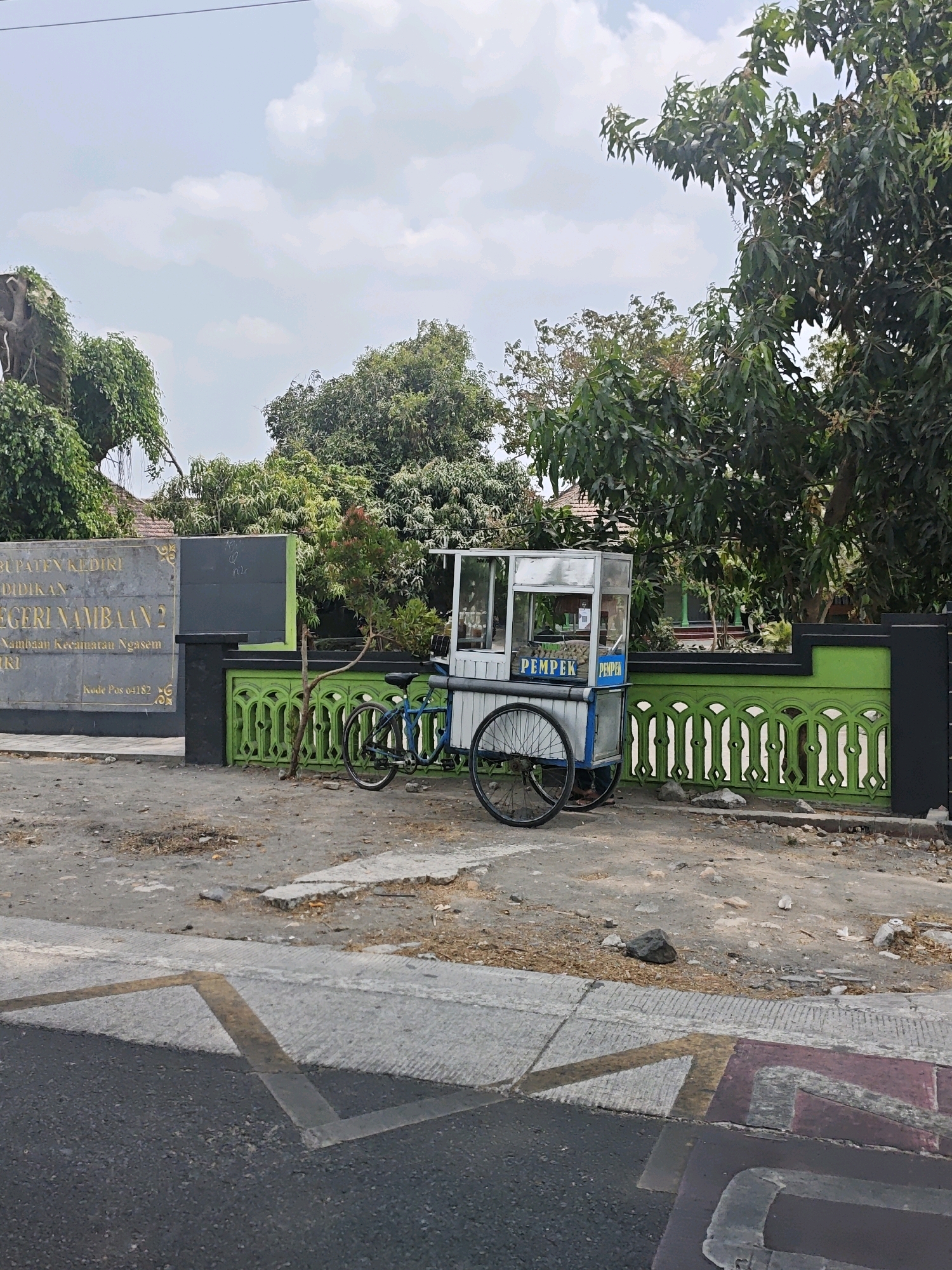 In Ngasem, Indonesia, a street scene is depicted. A food cart selling "PEMPEK" is parked on a dusty lot next to a green and black fence. The cart has a glass display case and a bicycle is attached to it. Behind the fence, a building with "PEMERINTAH KABUPATEN KEDIRI DINDIKAN NEGERI NAMBAAN 2" is visible, indicating this is likely the entrance to a school. Trees and vegetation surround the area, with a blue sky and scattered clouds overhead. The foreground shows a paved road with yellow and red markings, suggesting a crosswalk. The scene appears to be an outdoor commercial area in a semi-urban setting.