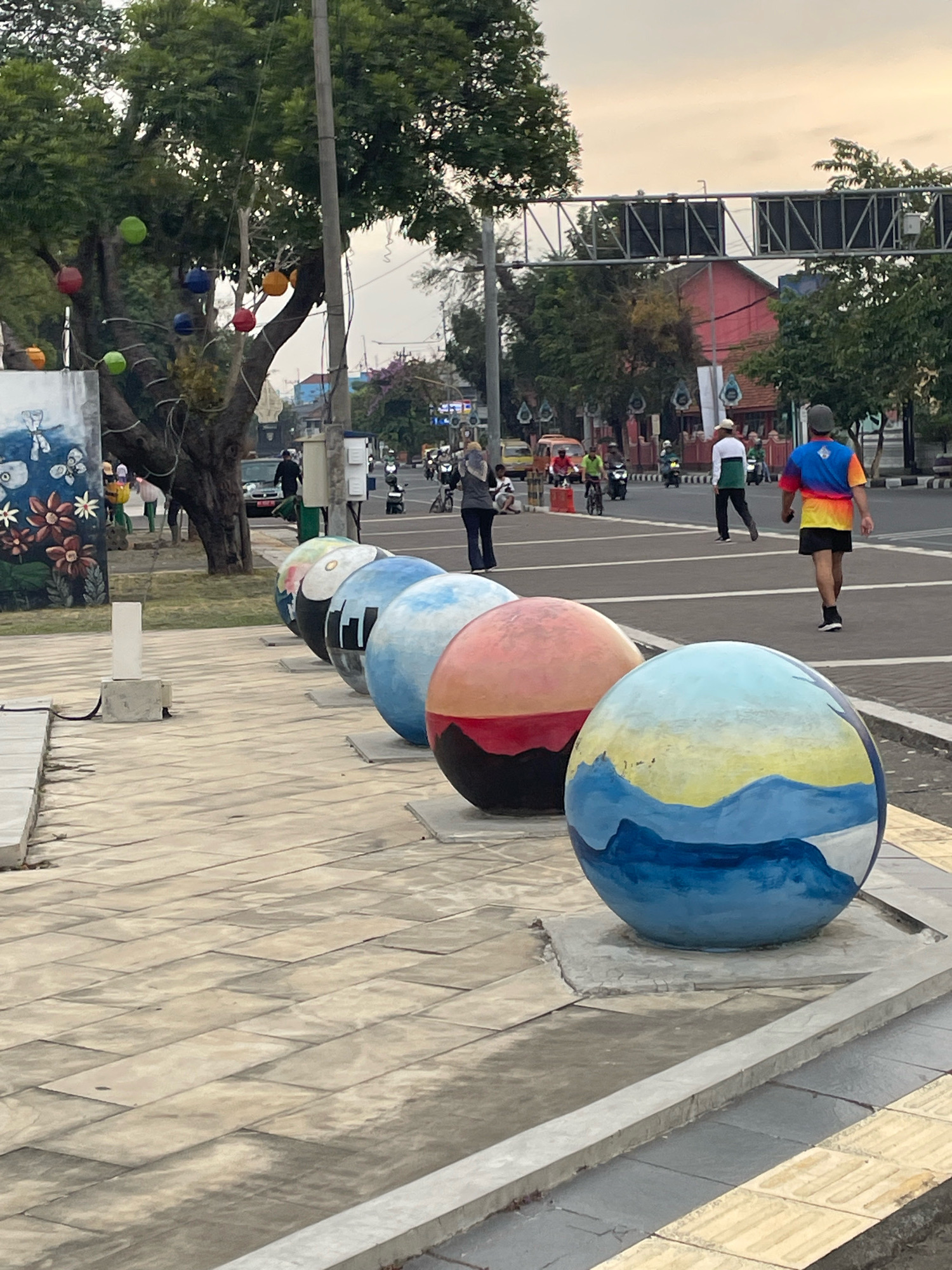 The image depicts a street scene in the city of Purworejo, Indonesia, near a decorative mural and several large, painted spheres. In the foreground, a row of these spheres, painted with various designs including a landscape with mountains and a sky, are positioned along a paved walkway. To the left, a mural featuring flowers and butterflies is visible on a wall. A large tree with colorful hanging lanterns stands behind the mural. In the background, a street with traffic, including motorcycles and cars, is apparent. Several individuals are walking or riding vehicles, and overhead, a traffic light structure is visible. The overall atmosphere suggests an urban setting with public art installations.
