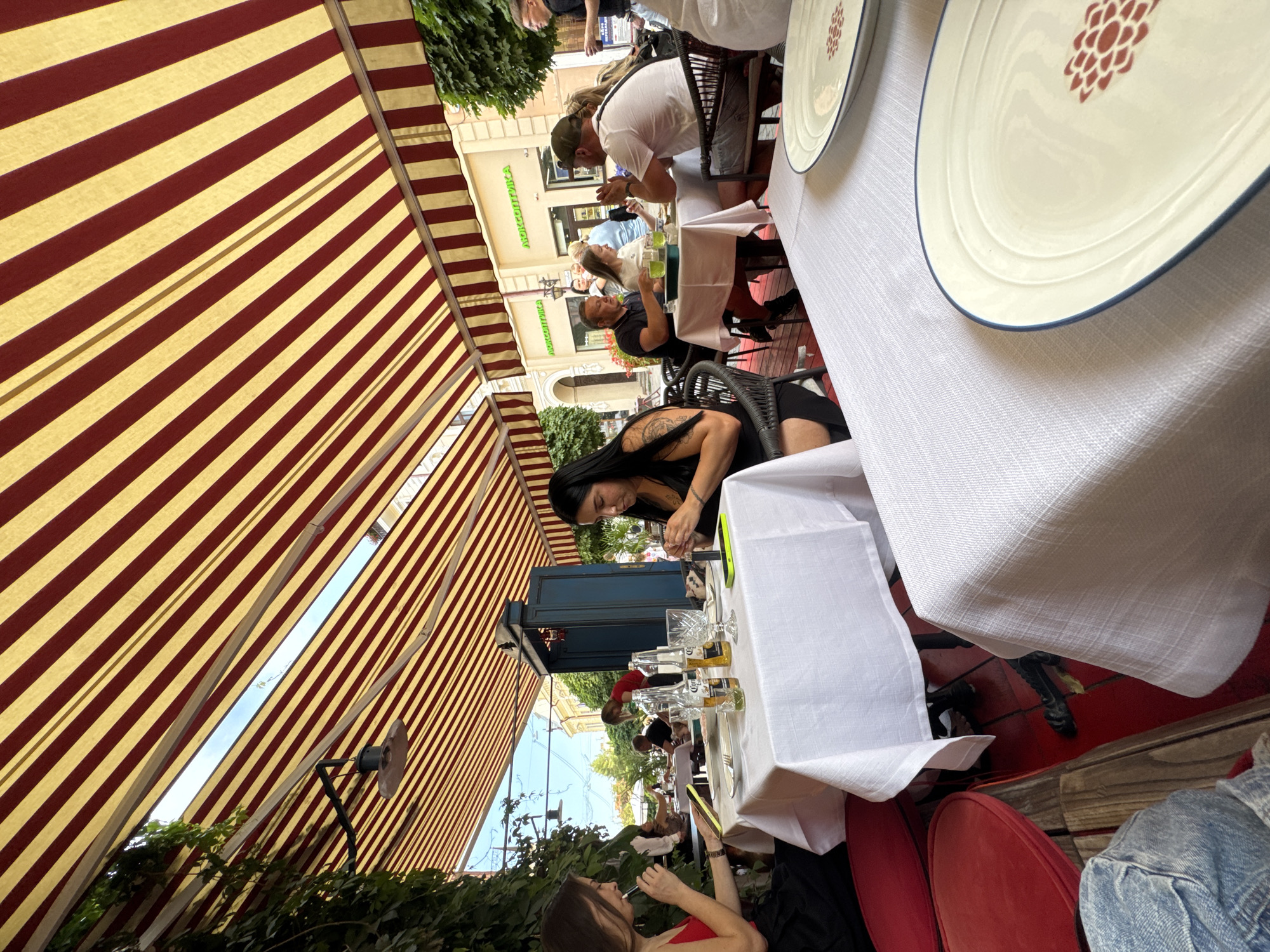 In a vibrant outdoor cafe setting in Chernivtsi, Ukraine, patrons are enjoying a meal under a striped awning of red and yellow. The scene is bustling with people engaged in conversation and dining. In the foreground, a woman with long dark hair and tattoos on her arm is seated at a table, looking down at her phone. Further back, a group of people are gathered around a table, some holding drinks and appearing to be in mid-conversation. The tables are covered with white tablecloths, and there are plates, glasses, and bottles of beer visible. In the background, buildings with balconies and greenery suggest a lively urban environment. The time of day appears to be daytime, with natural light filtering through the awning. The atmosphere seems relaxed and social. Visible text includes "АБХУДЖИКА" on a shop sign in the background.