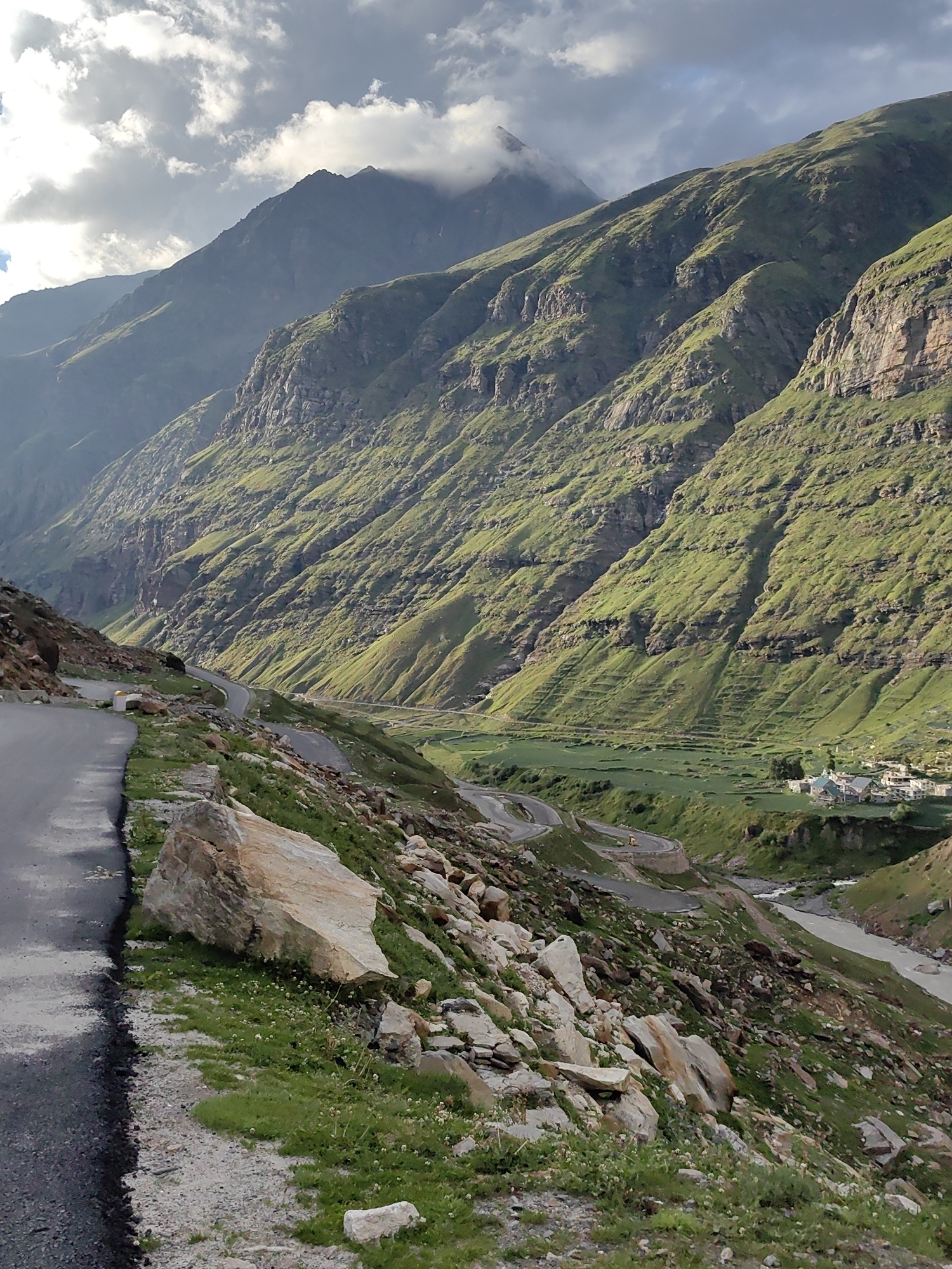 The image captures a sweeping vista of a mountainous region, likely the city of Yari-Khoksar, India, showcasing a dramatic landscape with a winding road and a distant village nestled in a valley. The foreground features a paved road curving to the left, bordered by a steep, rocky embankment covered in patches of green grass and scattered white wildflowers. Large, light-colored boulders are prominent along this embankment.
The mid-ground reveals the road continuing its serpentine path down into a lush green valley. This valley is characterized by terraced fields, suggesting agricultural activity. A river, appearing somewhat murky, flows through the valley floor, with a small cluster of buildings, a village, visible on the far bank. The architecture of the village appears to be traditional, with multi-story structures.
The background is dominated by immense, green-covered mountains that rise steeply from the valley. The slopes are rugged and etched with natural contours and some man-made terraces. The upper reaches of the mountains are partially obscured by dramatic, heavy clouds, with a peak visible piercing through the cloud cover. The lighting suggests it's either early morning or late afternoon, with the sun casting shadows and highlighting the textures of the mountainside. The overall atmosphere is one of rugged natural beauty and a remote, secluded settlement.