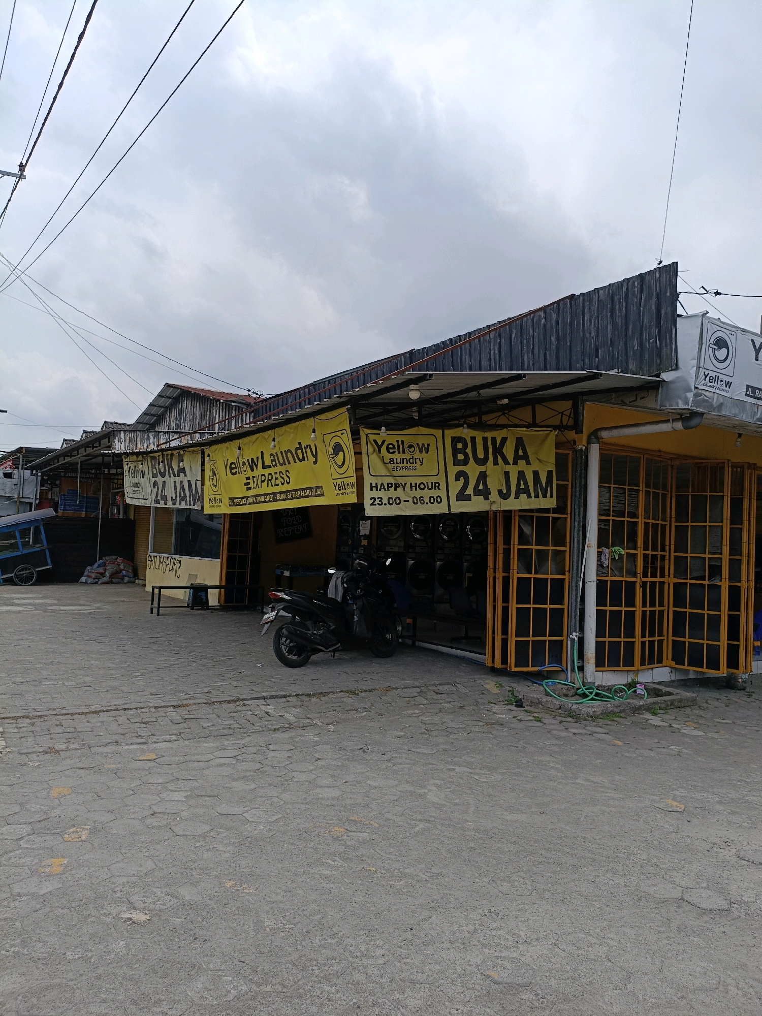 The image shows a street-level view of a laundry business in Maguwoharjo, Indonesia. The building is painted yellow and has large yellow banners that read "Yellow Laundry Express" and "Buka 24 Jam" (Open 24 Hours). There are multiple washing machines visible inside the establishment. A motorcycle is parked in front of the building. The pavement is made of interlocking concrete blocks. The sky is overcast.