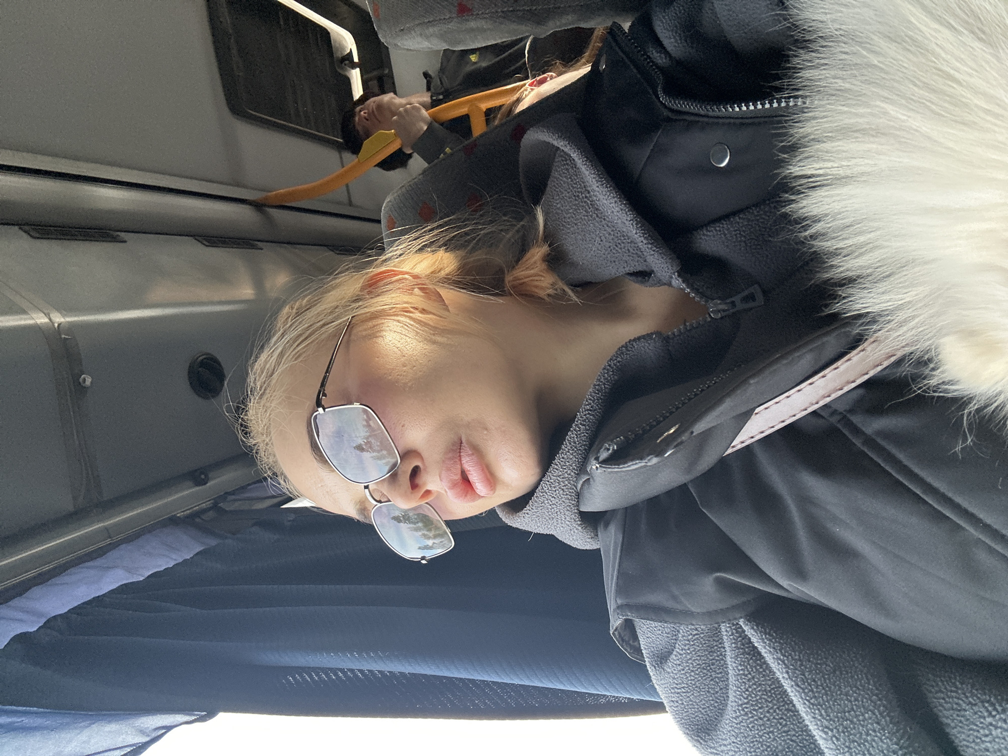 The image appears to be taken inside a bus, possibly in Megion, Russia. A young woman with light hair and sunglasses is visible, seemingly looking down. She is wearing a dark jacket with a white fur collar and a gray fleece underneath. A handbag strap is visible across her body. Another passenger is partially visible in the background, holding onto a yellow grab handle. The interior of the bus includes gray paneling, a window, and blue curtains.