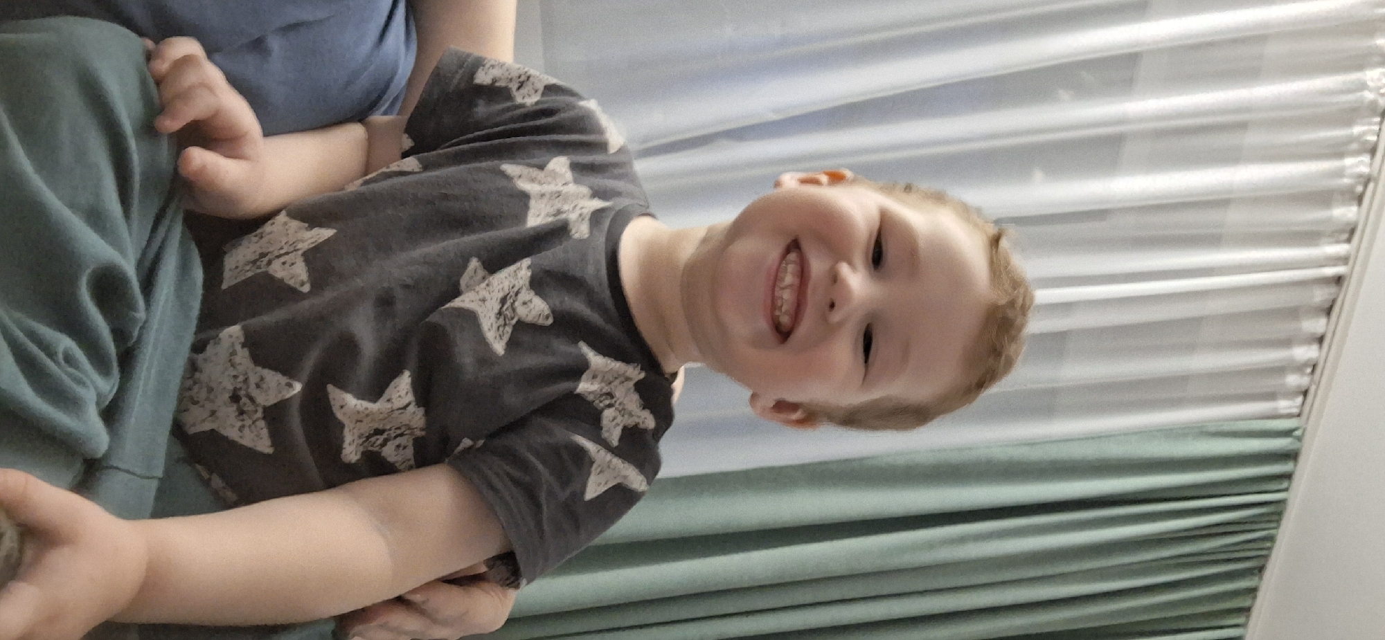 A young child, likely a boy with light brown hair, is captured in a close-up, rotated portrait, smiling widely and looking upwards towards the camera. He wears a dark gray t-shirt patterned with distressed light-colored stars. The child appears to be seated or held by an adult, with a plain blue-grey garment visible behind his upper body and teal or green fabric, possibly pants or a blanket, covering his legs and arms. His small hands are partially visible, one resting on the green fabric. The background features white horizontal window blinds or slatted panels in the upper part of the frame, and a pleated teal-green curtain or drape along the right side, indicating an indoor setting, likely during the daytime. The child's expression is joyful and happy, with an open-mouthed smile revealing his teeth. No text is visible.