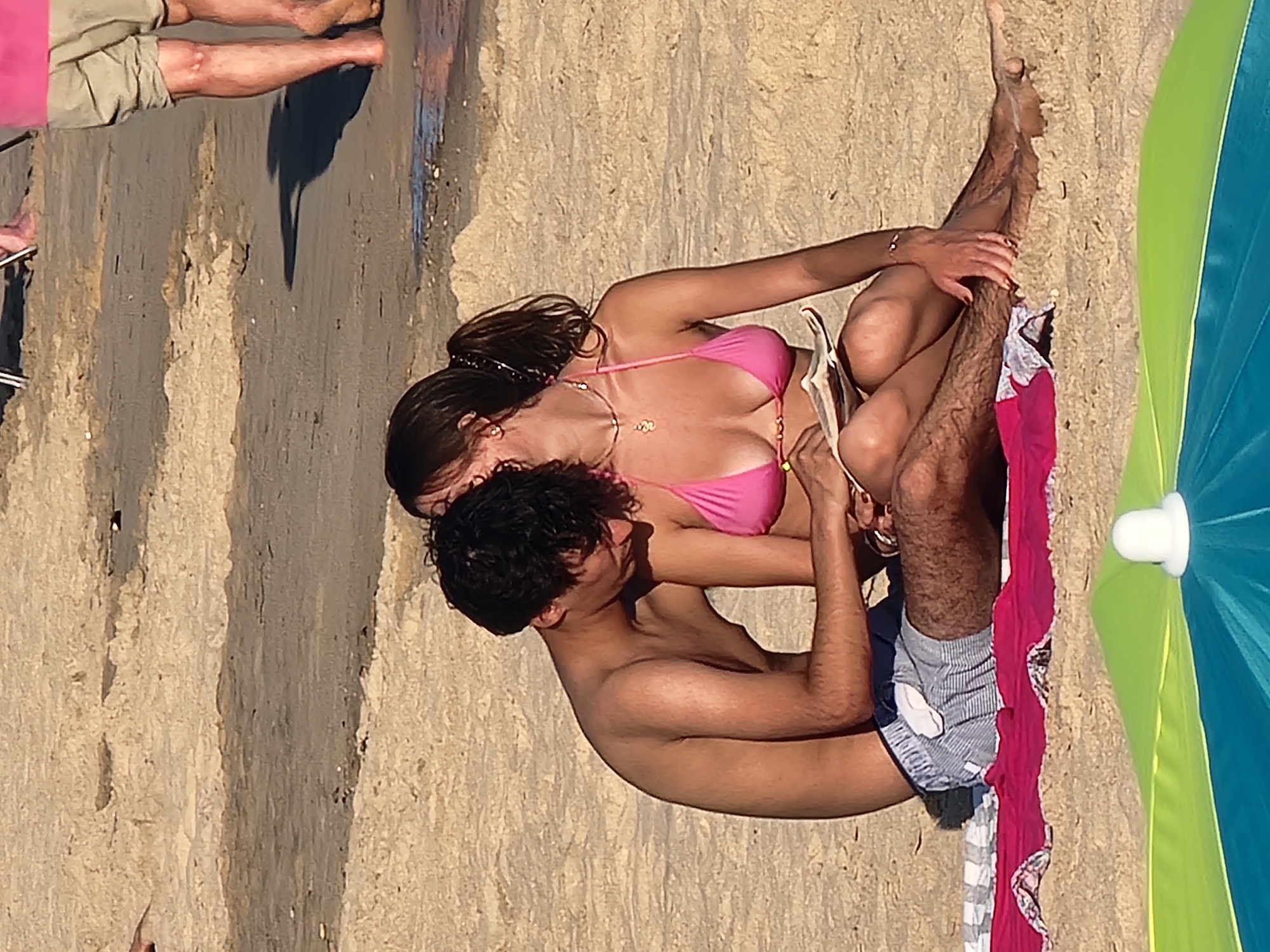 This image depicts a couple sitting on a sandy beach, likely in Quarteira, Portugal. The man, shirtless and wearing striped swim trunks, is seated on the left, leaning towards the woman. He has dark, curly hair and is looking down at something in his lap. The woman, on the right, is wearing a pink bikini and is also looking down, appearing to read or examine something held between them. Her dark hair is tied in a ponytail. They are positioned close together, suggesting an intimate interaction. The background shows the sandy beach with some slight undulations in the sand, indicating the natural terrain. In the foreground, a portion of a colorful beach umbrella is visible, its white central cap prominent against the blue and green fabric. The lighting suggests it's daytime, with the sun casting subtle shadows.