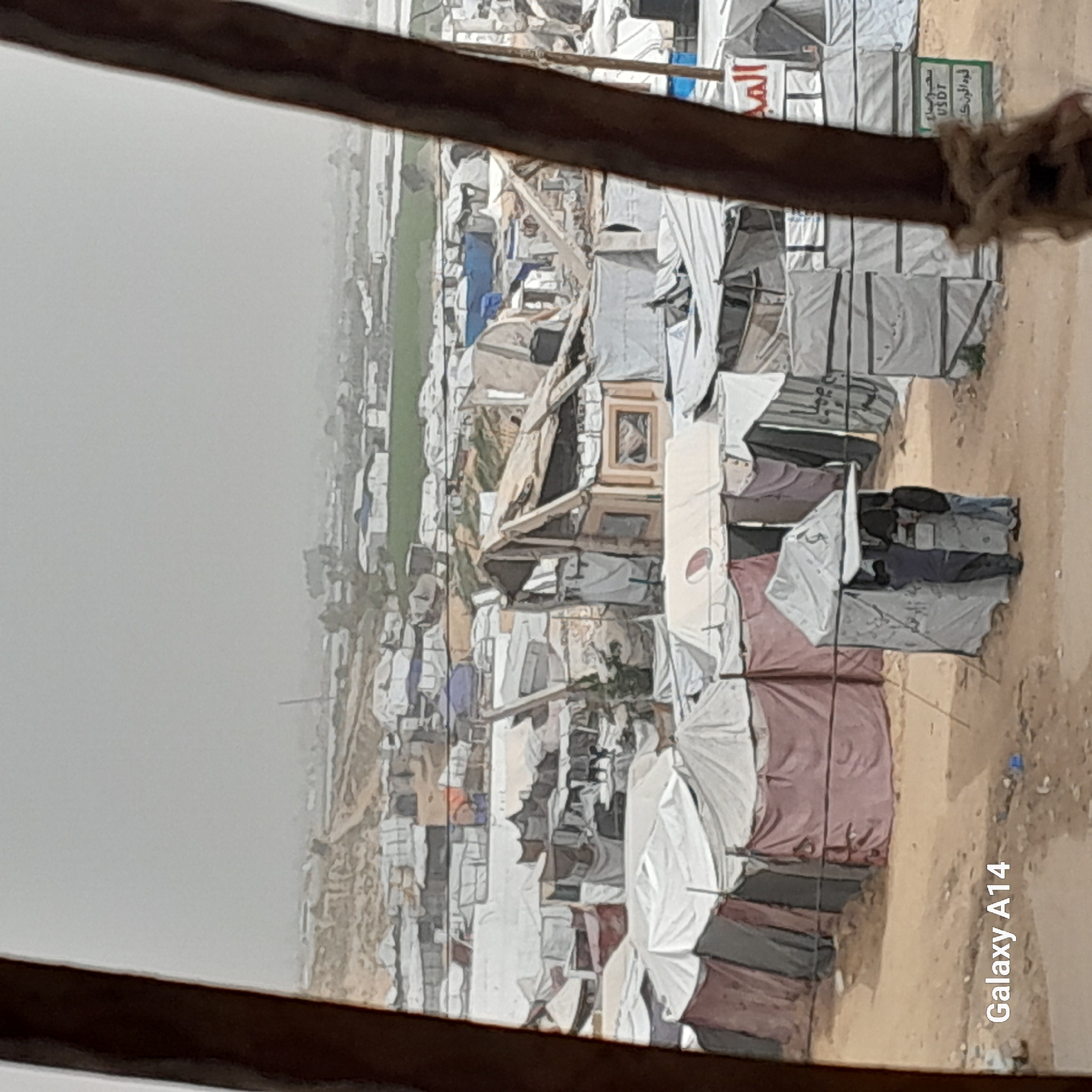 The image shows a densely packed refugee camp in Khan Yunis, Palestinian Territory. Numerous makeshift shelters, predominantly white and grey, are crowded together, made from various materials like fabric, corrugated metal, and wood. Some shelters appear damaged or collapsed. Open spaces are dusty. The sky is overcast, suggesting poor weather conditions. A sign with Arabic writing is visible. The scene conveys an atmosphere of displacement and deprivation.