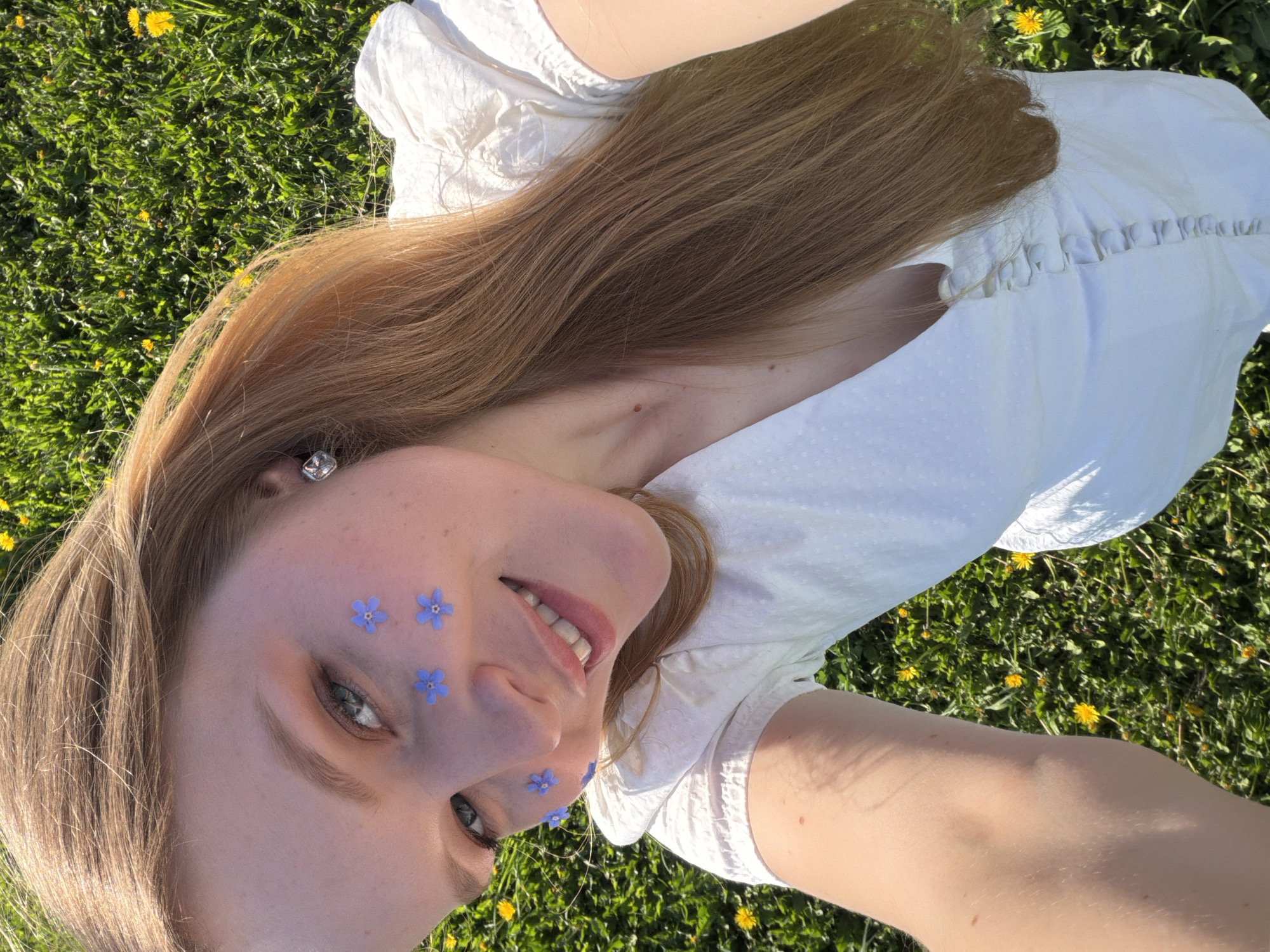 A young woman lies on her back in a grassy field dotted with dandelions. She wears a white dress and has long, brown hair. Blue flowers adorn her face. Her right arm stretches out towards the viewer, suggesting a selfie. Based on the image properties, the location is likely Кадряково, Russia, but there is no indication of city landmarks or architecture. The scene is outdoors, bathed in sunlight, suggesting a warm day.