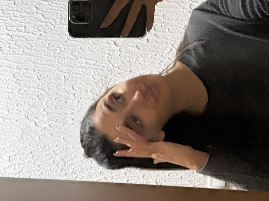 An image captured indoors in Zankovići, Montenegro, depicts a female subject taking a selfie in a mirror or reflective surface. The subject has dark hair, a light-to-medium complexion, and is wearing a dark, long-sleeved top with a crew neck. Her head is tilted slightly to the right, and her lips display a subtle, closed-mouth smile. Her right hand is raised, with fingers splayed, touching the right side of her head near the temple. Her left hand holds a dark smartphone, visible on the right side of the frame, indicating its use for capturing the image. The fingers of the left hand show light-colored nail polish. The background primarily consists of a textured white wall. A vertical strip of dark wood and a partially visible white electrical outlet or switch are present on the far left edge of the frame.