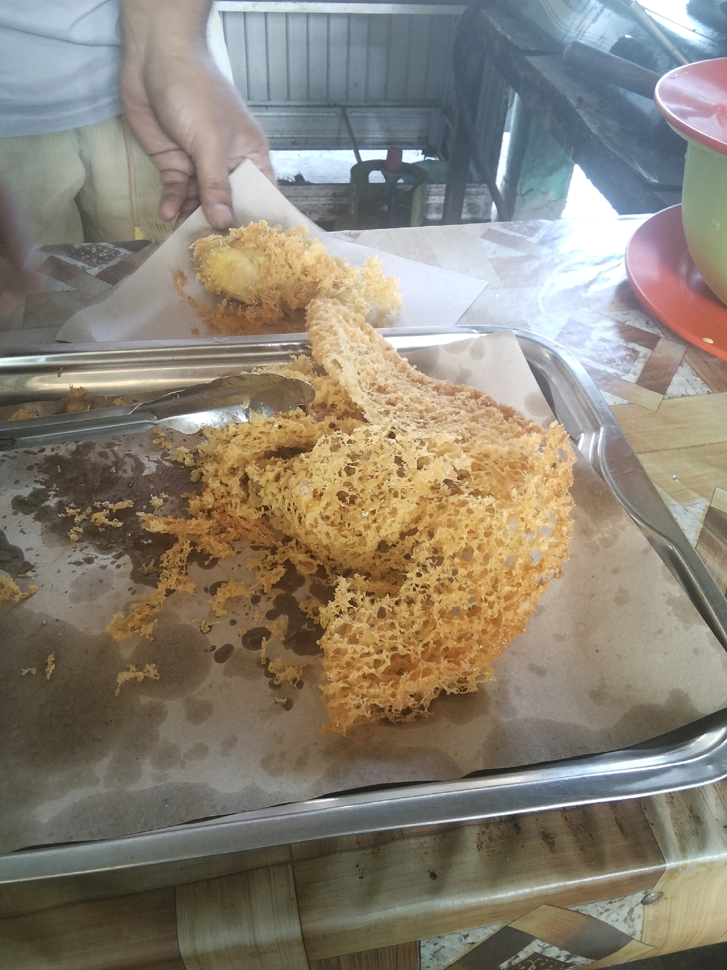 In Magelang, Indonesia, at a street-side food stall, a vendor is serving freshly fried snacks. The snacks, which appear to be a type of crispy fritter, are golden brown and piled on a metal tray lined with brown paper. A pair of tongs rests on the tray, having just been used to handle the food. In the background, a red and green bowl sits on a patterned table, and a person's hands, clad in a light-colored shirt and beige pants, are visible as they hold up a piece of the fried snack, preparing to place it on the tray. The environment suggests an outdoor dining or preparation area, common in local Indonesian food establishments.
