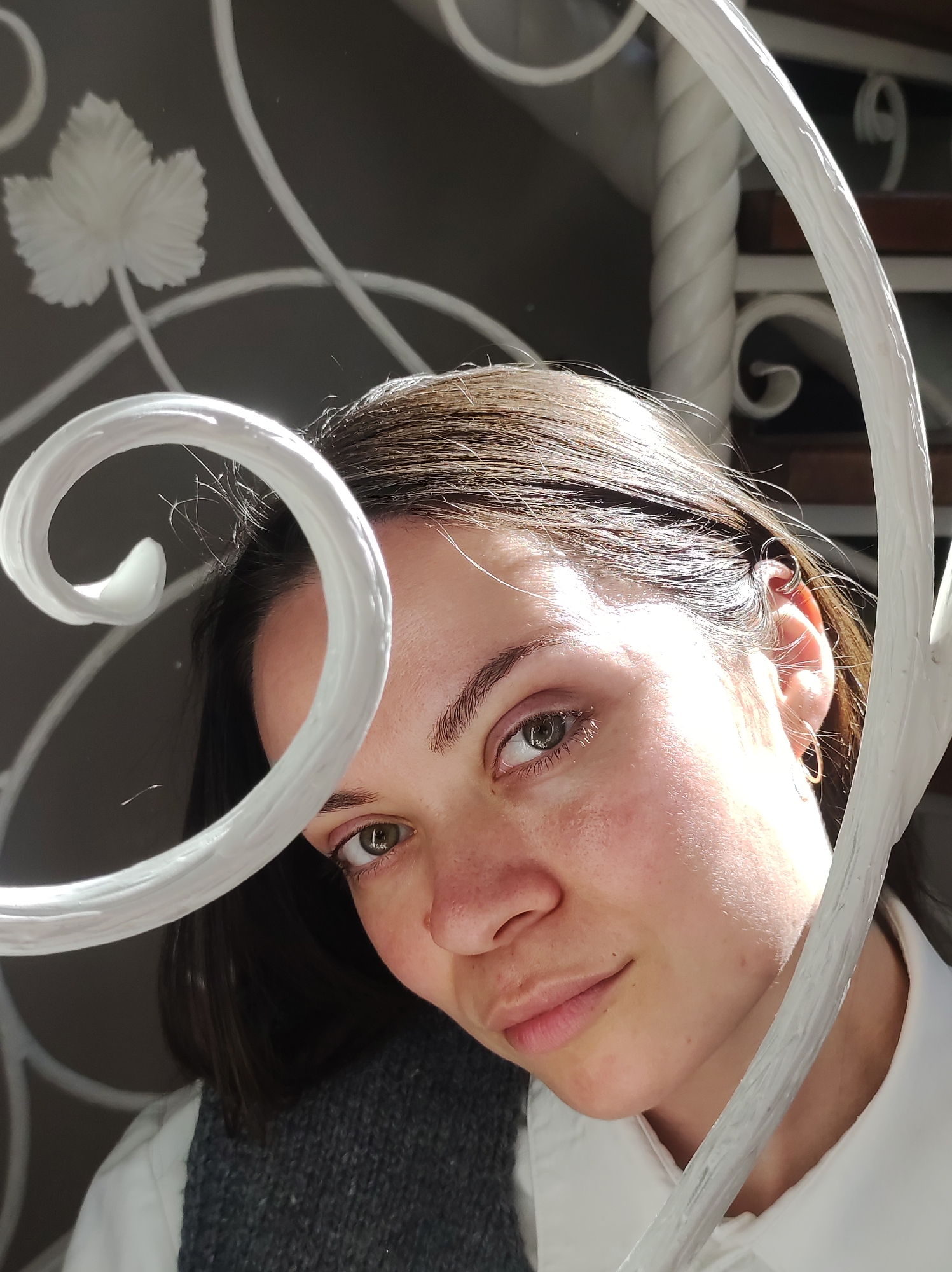 A close-up portrait of a woman with fair skin, light brown hair, and hazel eyes. She is positioned behind a decorative white metal railing with spiraling elements. She is wearing a white shirt with a gray vest visible beneath. The background is slightly out of focus, showing hints of a wall with a decorative white leaf design and a portion of a white spiral staircase. The woman's gaze is directed towards the camera, creating direct eye contact. The lighting is bright and somewhat harsh, casting shadows on her face and highlighting the texture of the metal railing. No information regarding the location is available.