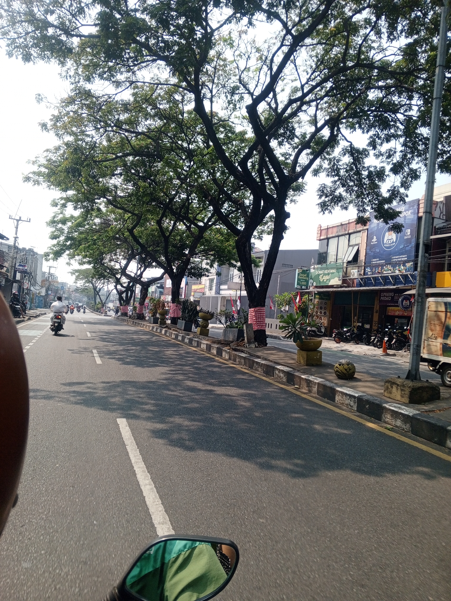 This is a street-level, medium-shot, eye-level photograph taken from the perspective of a motorcycle rider in Depok, Indonesia. The scene depicts a moderately busy street lined with trees on the left side, casting dappled shadows across the asphalt road. On the right side of the road, there are several commercial buildings, some of which have awnings and signage. One building prominently features a large banner for "EXP". The street is paved with asphalt and has a white dashed line marking the lane. Several motorcycles are visible on the road, with one rider in the foreground on the left side of the lane, heading away from the camera. The sky is bright and appears to be daytime. The overall impression is a typical urban street scene in Indonesia, with elements of commerce, transportation, and natural foliage.