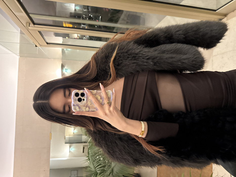 A woman takes a selfie in a mirror. She wears a brown, form-fitting dress with sheer panels. Over it, she drapes a fur stole, gray on one side and black on the other. Her hair is long, dark, with caramel highlights. She holds up a phone with a textured, decorative case. Her nails are long and painted a light color. A gold bracelet is visible on her wrist. The background appears to be a well-lit, modern indoor space with large windows.