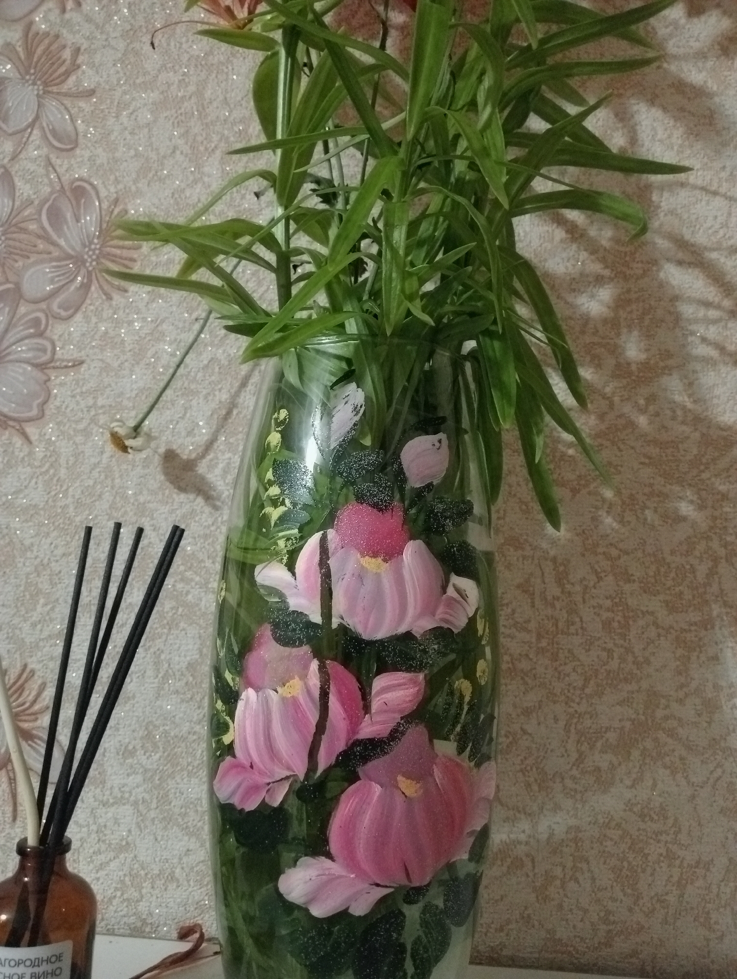 In a domestic indoor setting, a tall, clear glass vase stands on a surface, filled with greenery and artificial pink flowers. The vase is decorated with painted pink flowers and dark green foliage, some of which has a glittery finish. The background is a textured, light-colored wallpaper adorned with a subtle floral pattern. To the left of the vase, a small brown glass bottle with a label containing Cyrillic text is visible, along with a set of dark reed diffusers. The image does not depict any people or specific city landmarks.