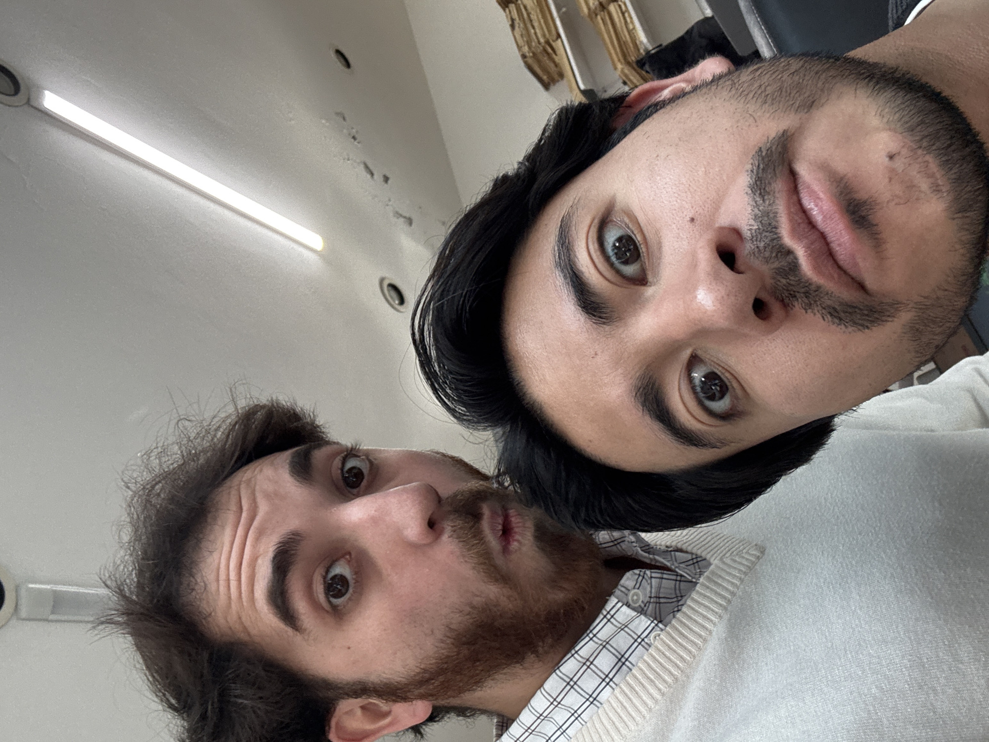 Two men are posing closely for a selfie indoors. The man on the right has dark hair, a mustache, and a beard, and wears a light-colored sweater. The man on the left has shorter, disheveled dark hair and a beard, and is wearing a checkered shirt and light-colored sweater. The background includes a bright fluorescent light, a white wall, and some partially visible items that could be clothes hangers. The photograph does not contain sufficient visual information to determine whether it was taken in Puebla, Mexico.