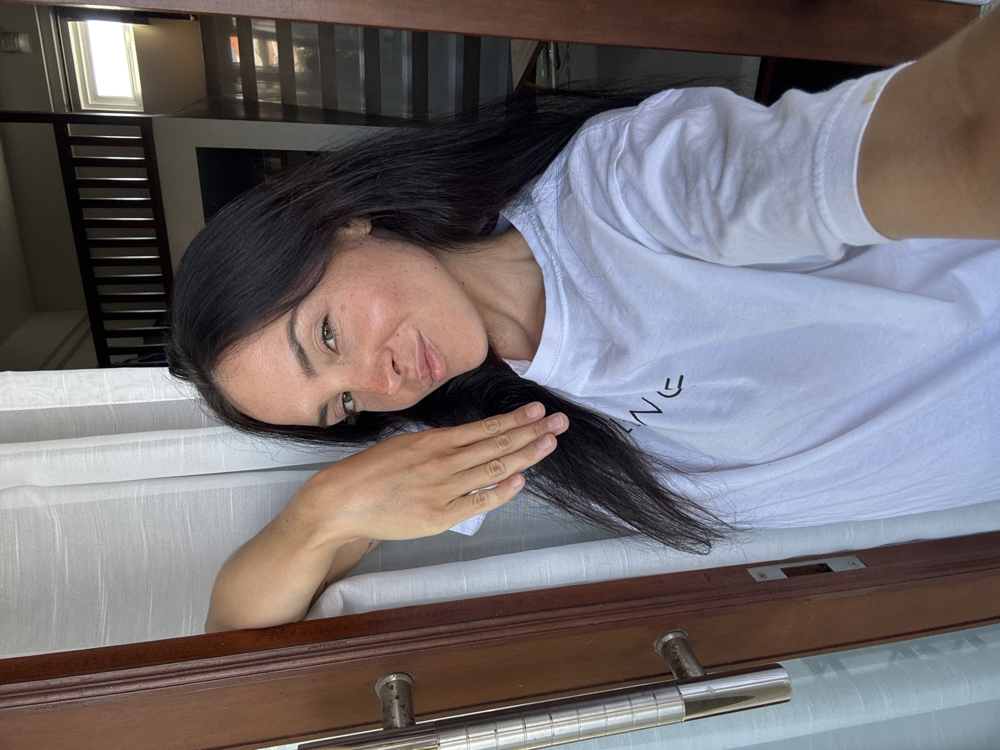 A young woman with long, dark hair and fair skin is leaning on a wooden doorframe, partially obscured by a white curtain. She is wearing a white t-shirt with black lettering. Her right arm is bent at the elbow, with her hand resting on her head. Her left arm is bent and extended downwards, with her hand resting on the doorframe. Her gaze is directed towards the camera, and she has a slight smile. The background appears to be an indoor setting with wooden railings and a window. The lighting suggests it is daytime. The location is identified as North Kuta, Indonesia.