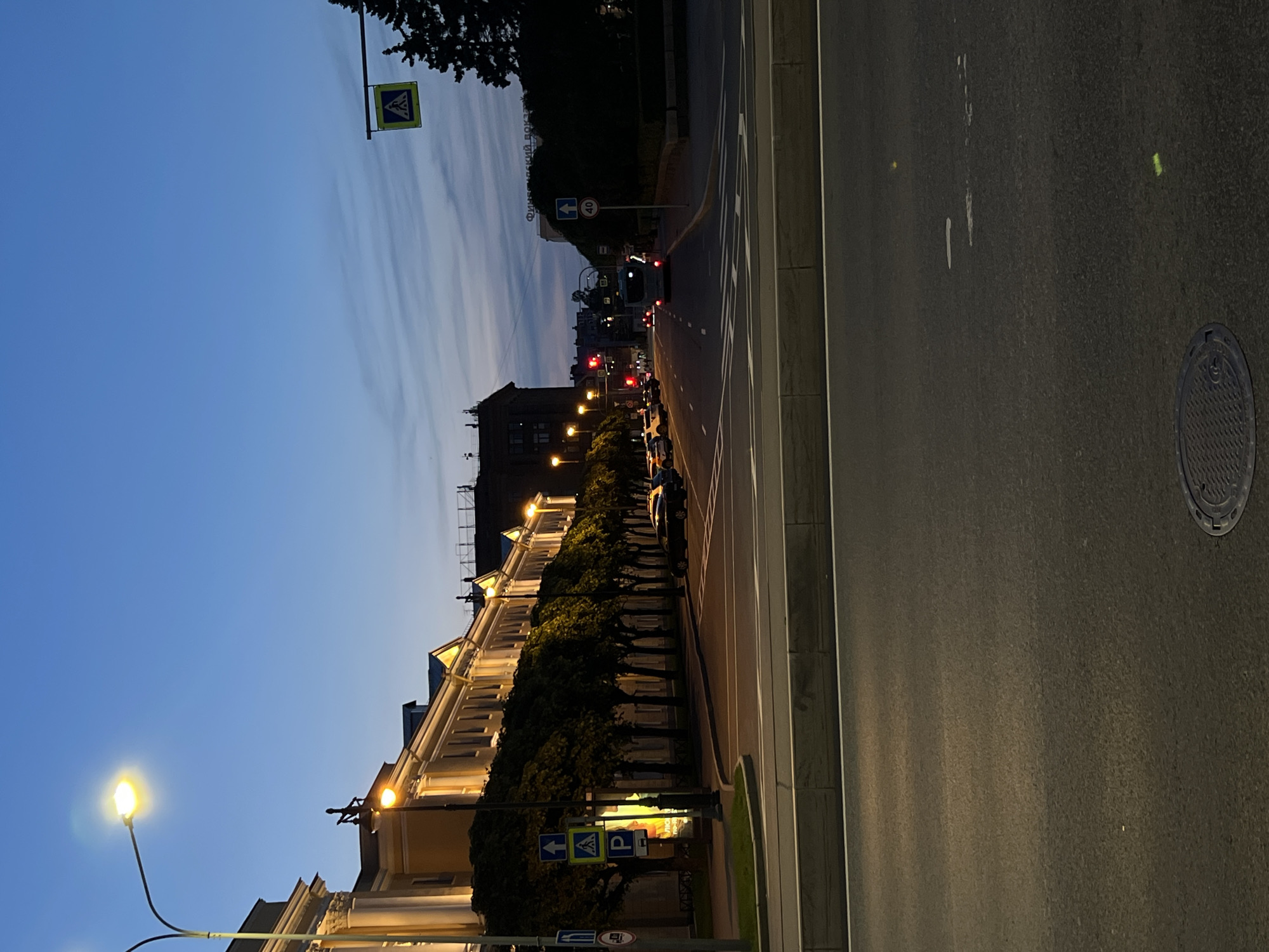 The image depicts a street scene at dusk in an urban environment, likely in Russia given the visible signage. In the foreground, a paved road occupies the lower half of the frame, featuring white lane markings and a manhole cover. The middle ground shows a street lined with trees and buildings. Several cars are parked or driving on the street. Traffic lights are visible, with one showing red. A pedestrian crossing sign is present. The background reveals more buildings and the sky, which is a deep blue with wispy clouds, indicating twilight. The overall atmosphere is calm and quiet, with streetlights and building lights illuminating the scene.