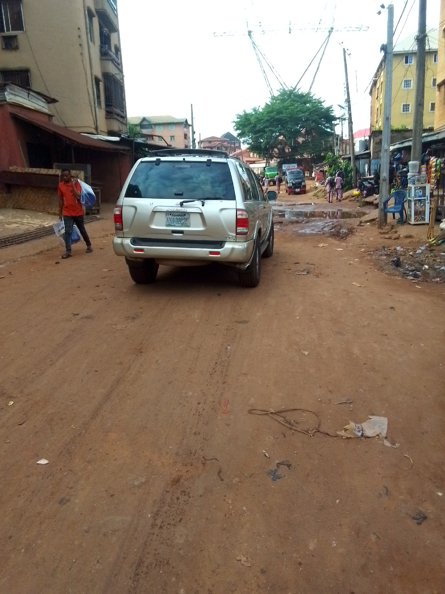 The image depicts a street in Onitsha, Nigeria. The unpaved road is primarily reddish-brown soil and is muddy. A silver SUV, oriented away from the viewer, is parked on the road. A man wearing an orange shirt and carrying a blue bag is walking along the left side of the vehicle. In the background, there is pedestrian and vehicular traffic. There are buildings of varying heights on both sides of the road. Electrical poles and wires are visible overhead. Debris and litter are scattered on the road surface.