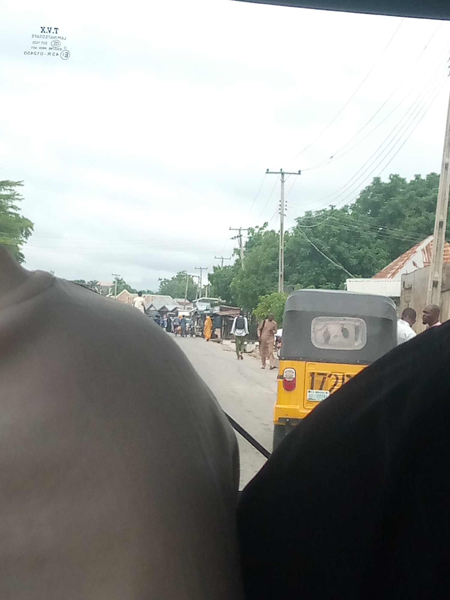 The image depicts a street scene in Jalingo, Nigeria, as seen from inside a vehicle. In the foreground, a large portion of a person's shoulder and upper arm, covered by a black garment, is visible on the right side. On the left side, another person's shoulder and upper back, with light-colored skin, occupy the lower left quadrant.
The middle ground shows a yellow auto-rickshaw, a common mode of transport in Nigeria, with its rear license plate clearly visible displaying "172". The auto-rickshaw has a gray canopy. Several people are visible walking or standing on the street and the sidewalk. One individual in a light brown robe is walking away from the camera. Another person wearing a white shirt and dark pants with a white backpack is walking towards the camera. Further down the street, a group of people are gathered, some appearing to be engaged in conversation.
The background reveals a typical street in Jalingo with buildings that appear to be low-rise structures with corrugated iron roofs, characteristic of many urban areas in Nigeria. Electrical poles and wires are strung along the street, indicating the presence of electricity infrastructure. Trees with lush green foliage line the sides of the street, suggesting a tropical or sub-tropical climate. The sky is overcast, indicating a cloudy day. The overall impression is a bustling street scene in a Nigerian city.