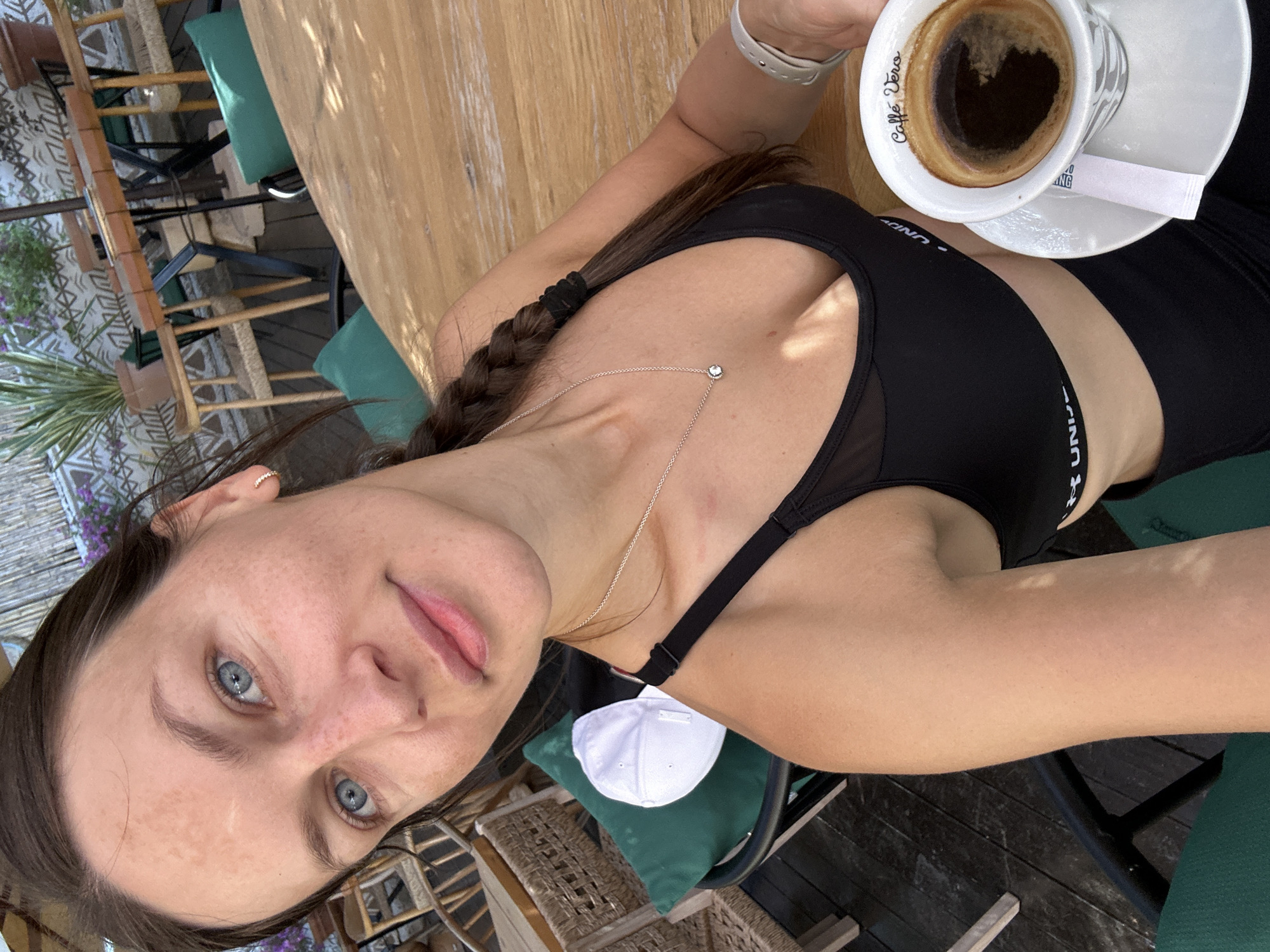 A young woman with blue eyes and light skin is taking a selfie at an outdoor cafe. She has brown hair in a braid and is wearing a black sports bra. She is holding a cup of coffee with a heart-shaped crema. The table is made of wood, and the background shows outdoor seating with green cushions and wicker furniture. There is also a white baseball cap visible. The lighting suggests it is daytime, and the overall mood is relaxed and casual. The text "Caffè Vero" is visible on the coffee cup. The woman appears to be enjoying her coffee in a pleasant outdoor setting.
