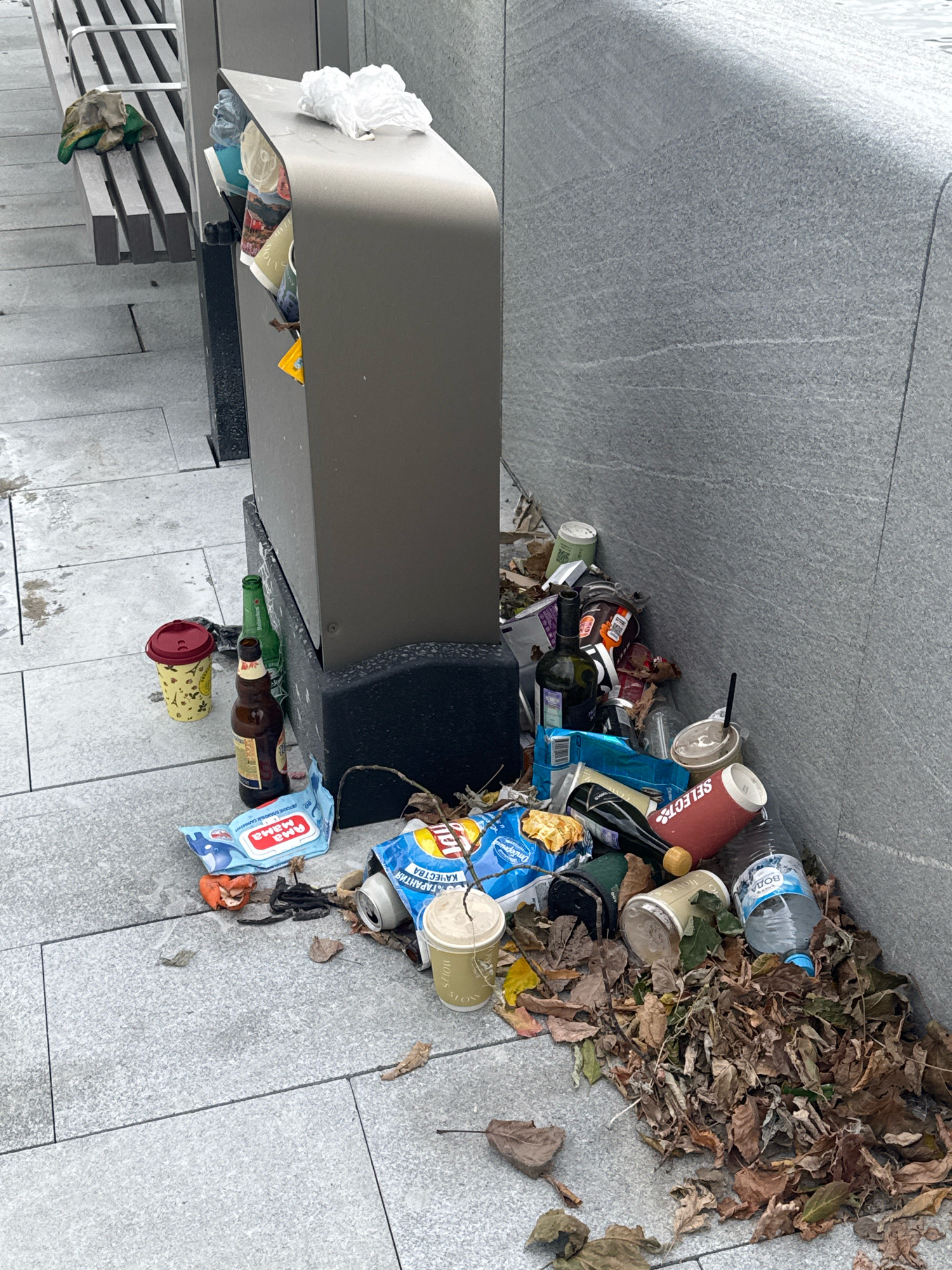 An outdoor public space in Moscow, Russia, features a modern trash receptacle overflowing with litter, including empty beverage containers, food wrappers, and leaves. A portion of a metal bench with some discarded gloves is visible to the left. The ground is paved with large gray tiles, some of which are wet. The wall to the right appears to be made of textured gray stone.