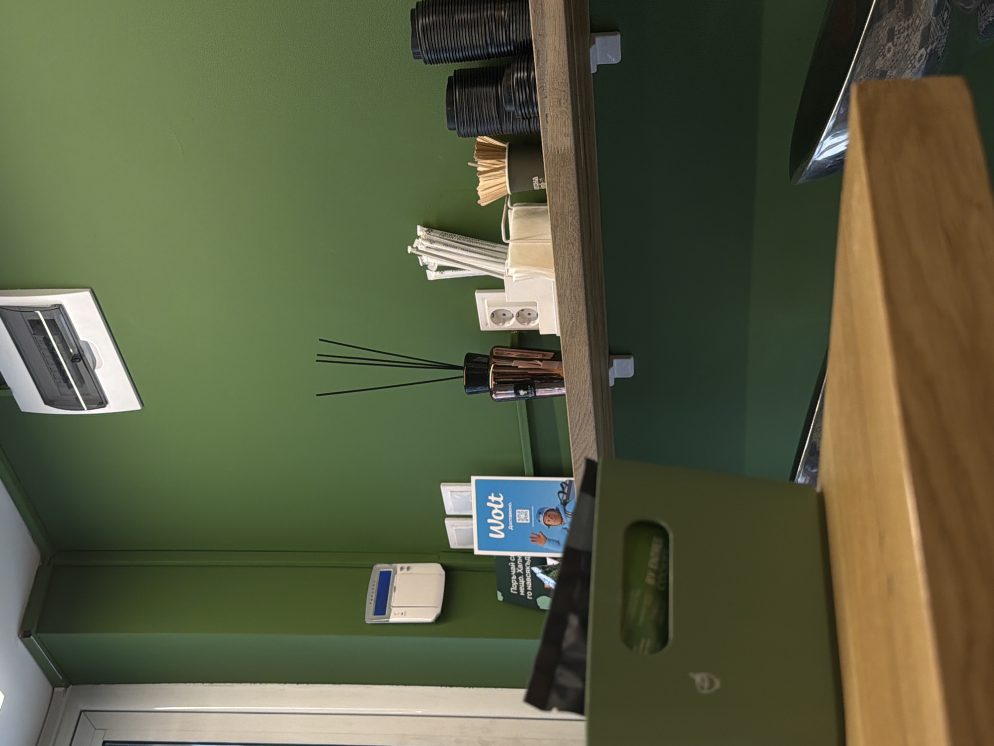 The image depicts a retail or service environment, likely a cafe or a small shop, with a prominent green wall as the backdrop. On the wall, at the top left, a white air conditioning unit is mounted. To the right of the air conditioner, a white electrical panel with multiple switches is installed. Below this, a white device with a blue display, possibly a security system panel, is visible.
In the center of the frame, a wooden shelf runs horizontally. On this shelf, several items are arranged. From left to right, there is a dark copper-colored diffuser bottle with black reeds. Next to it, a white dispenser holds coffee stirrers. To the right of the dispenser, a stack of white paper cups is visible, along with a white box containing what appears to be sugar packets or creamers. Further to the right, a dark gray container holds more coffee stirrers. The rightmost items on the shelf are stacks of black disposable cups, some with lids.
In the foreground, a green container with handles sits on a wooden surface. The container has a logo or text printed on it, which is partially visible. A wooden table or counter is also in the foreground, out of focus.
The overall scene suggests a functional area where amenities for customers or staff are provided. The presence of Wolt branding on a blue sign on the shelf indicates a partnership or service associated with food delivery. The location is identified as Sofia, Bulgaria.