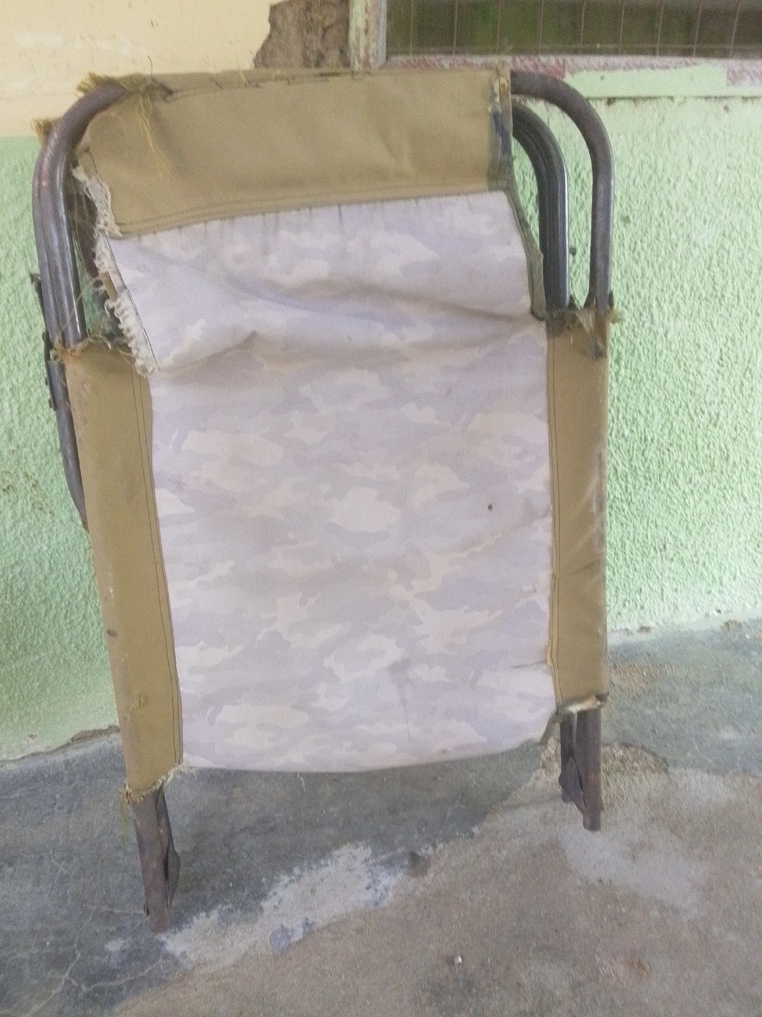 This media file is a medium-shot, eye-level photograph of a damaged, foldable cot lying on a concrete floor. The cot has a metal frame with exposed, rusted metal legs. The fabric of the cot is a light grey camouflage pattern, with a tan fabric lining along the edges. The fabric appears worn and frayed in places, suggesting age and disuse.
The cot is leaning against a textured, light green wall. Above the cot, a section of a light brown, rough wall is visible, with some damage and peeling paint. A metal grate is visible at the very top of the image, suggesting the cot might be in an outdoor or semi-enclosed area. The floor beneath the cot is a rough concrete surface with some discoloration and cracks.
The lighting appears to be natural, suggesting daytime, and there are no strong shadows. The overall impression is one of neglect and disrepair. The setting might be an outdoor storage area, a makeshift shelter, or a place where such equipment is kept when not in use, given the rough walls and floor. No people are visible in the image. There is no visible text. The location context of Jalingo, Nigeria, suggests a potentially warm climate, though the image itself does not provide specific weather cues.
