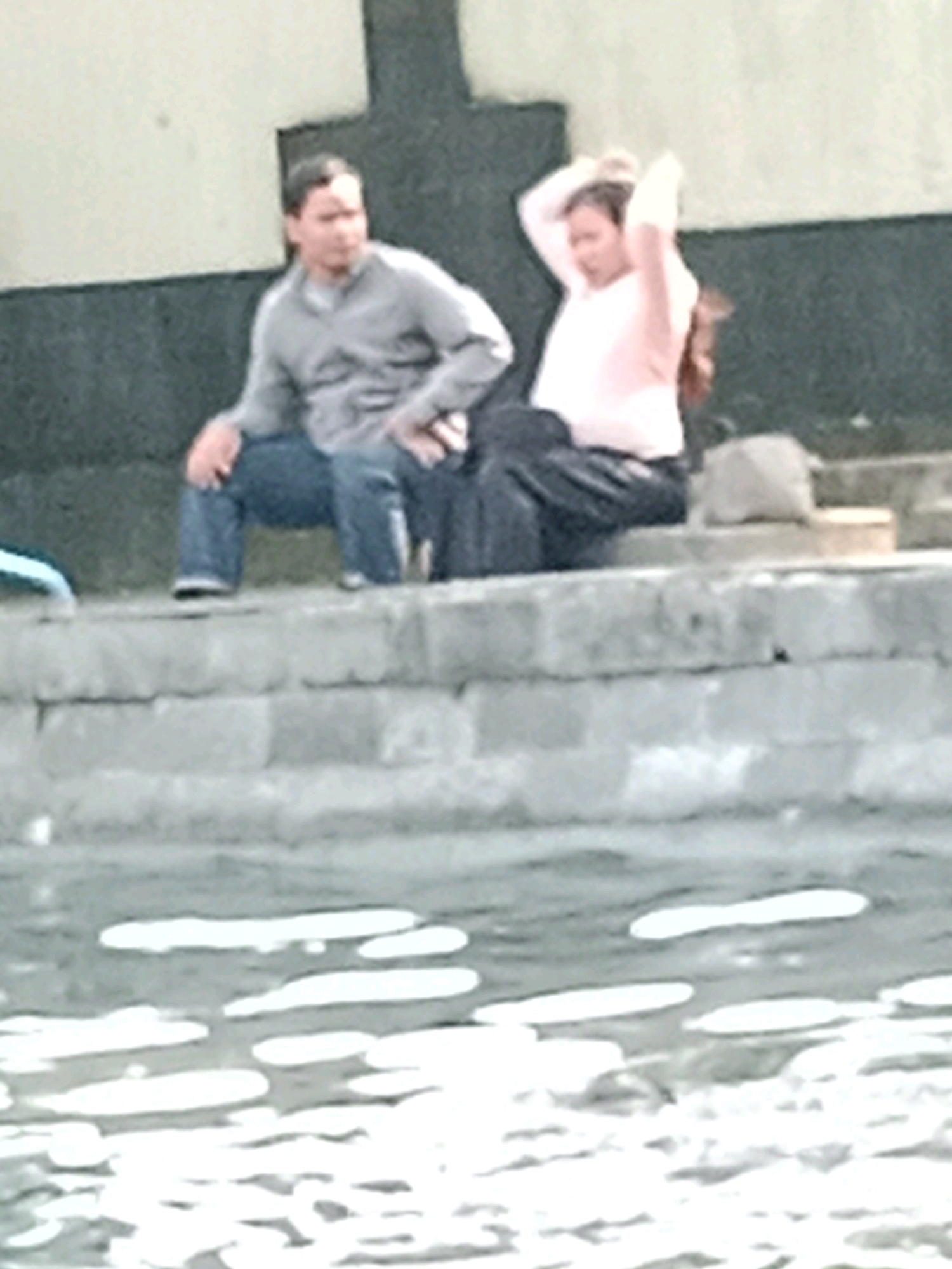 In Jentekan, Indonesia, two individuals are seated on a stone ledge overlooking a body of water. On the left, a male wearing a gray collared shirt and blue jeans is seated with his legs spread. To his right, a female wearing a light pink long-sleeved shirt and dark pants sits with her legs bent, her hands raised to her hair, and her head tilted back. The water in the foreground reflects light, creating abstract shapes. The background features a textured, light-colored wall.