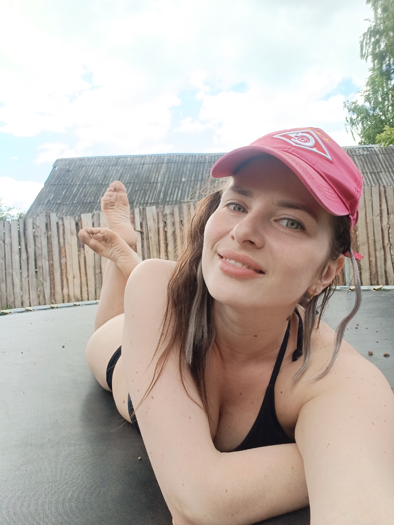 A woman is lying on a trampoline with her upper body angled towards the camera. She is wearing a black bikini top and a pink baseball cap. Her hair is brown and appears wet, with some strands falling around her face and shoulders. The background shows a rustic wooden fence and a building with a corrugated roof, possibly in a rural setting. The sky is partly cloudy. Her feet are visible in the upper left portion of the image, propped up and crossed. The woman has green eyes and is smiling. The overall impression is a casual, outdoor scene. There is no specific indication of the location being in Rusalevka, Russia.