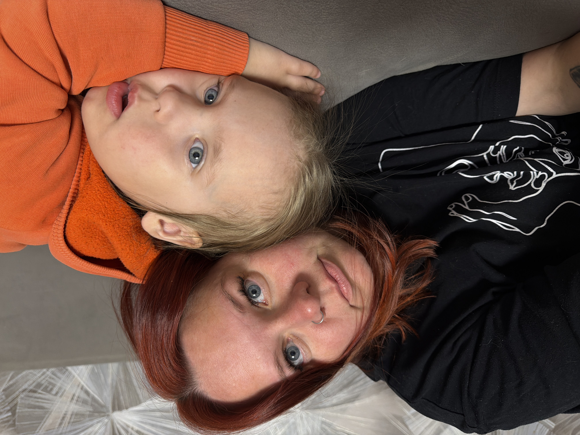 A mother and child lie on what appears to be a bed or soft surface. The child, wearing an orange hooded garment, is positioned at the top. The adult woman with red hair is positioned at the bottom, wearing a black shirt adorned with white line drawings. Both individuals are looking directly at the viewer. The location in the city of Chaikovsky, Russia is not discernible from the image.