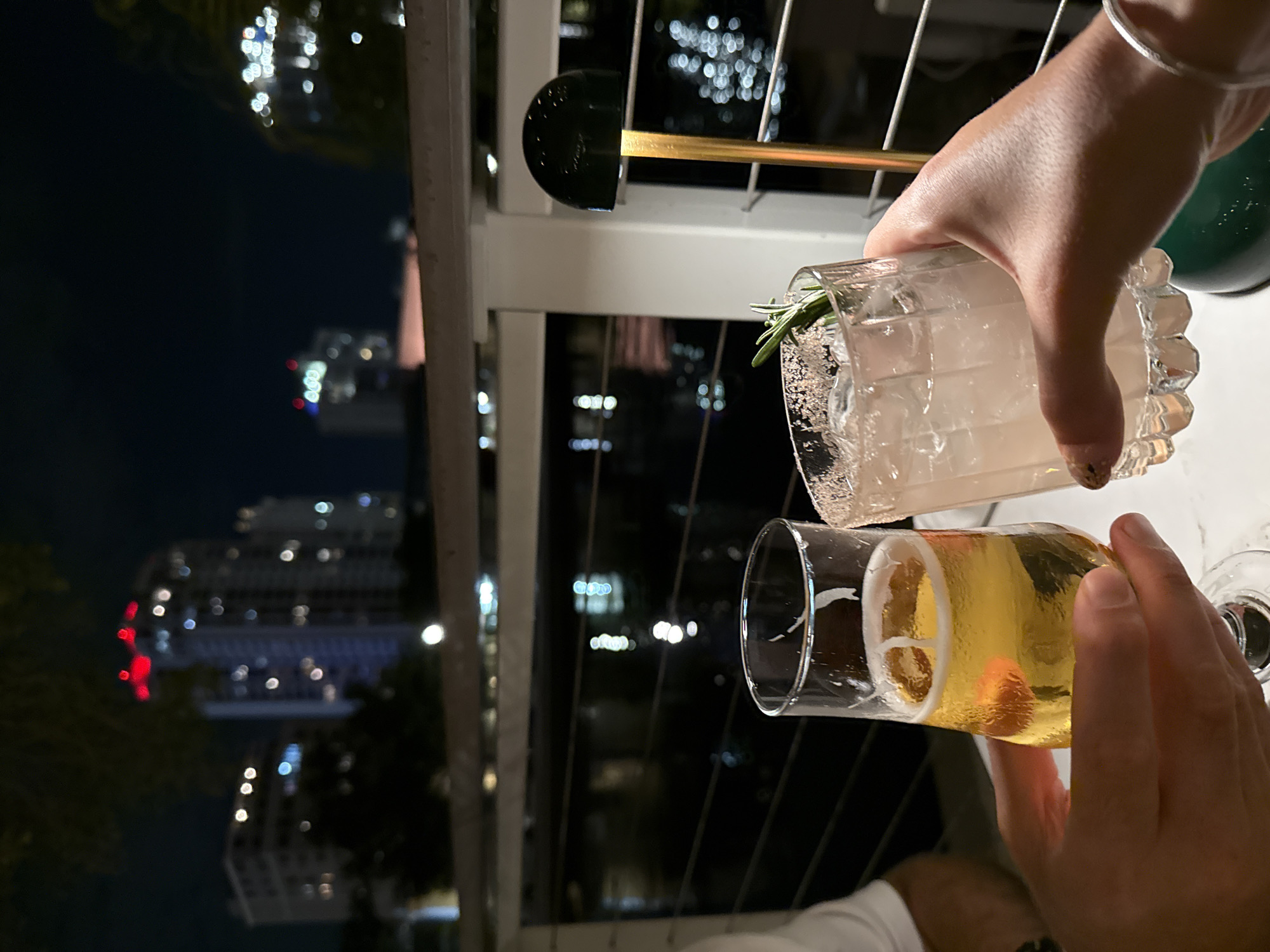 The image depicts a nighttime outdoor scene in Saint Petersburg, United States of America, likely at a waterfront establishment. Two individuals are engaged in a toast, holding alcoholic beverages. The person on the left holds a clear glass containing golden-yellow beer with a foam head. The person on the right holds a faceted clear lowball glass containing a light pink or cloudy cocktail with ice, garnished with a sprig of rosemary, and featuring a salt-rimmed edge. Both sets of hands are visible holding their respective glasses over a light-colored table surface.
In the mid-ground, a white railing system with horizontal bars separates the immediate foreground from a dark body of water, which reflects blurred lights from the city. In the background, several multi-story buildings of the city skyline are visible, illuminated by various internal and external lights, with some distant rooftops exhibiting red lights. Dark foliage is present between the water and the buildings. A dark green, dome-shaped portable table lamp with a gold-colored stem is partially visible on the right side of the image, behind the cocktail glass. The overall environment is dark, illuminated primarily by artificial lights from the city and the establishment.