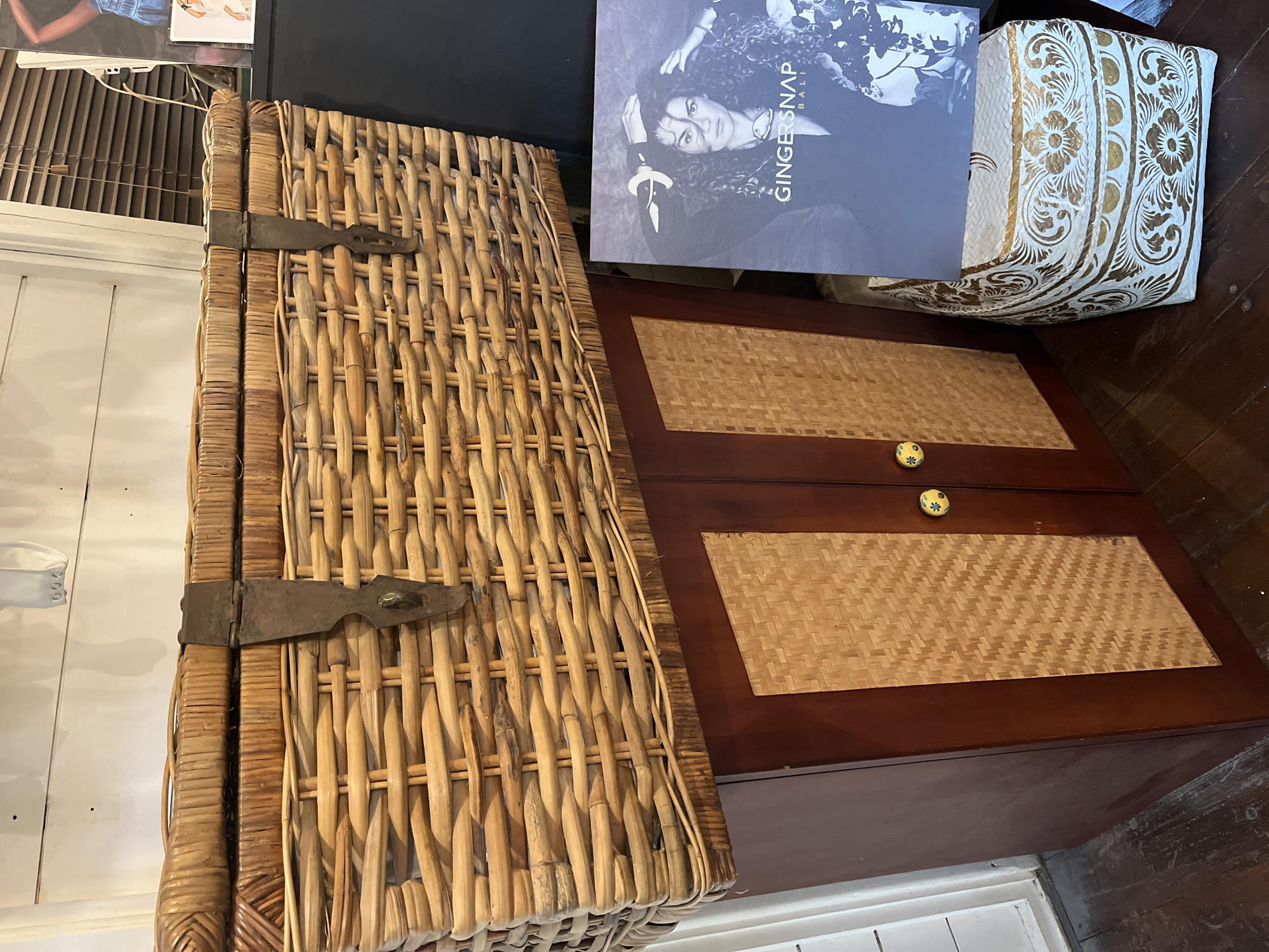 A large, woven wicker trunk with metal clasps sits atop a dark wooden cabinet with two woven panels and decorative yellow knobs. To the right of the cabinet is a framed picture of a woman and a small decorative woven basket with a gold floral pattern. The setting appears to be an indoor retail space, possibly in North Kuta, Indonesia, given the presence of items commonly found in Balinese shops.