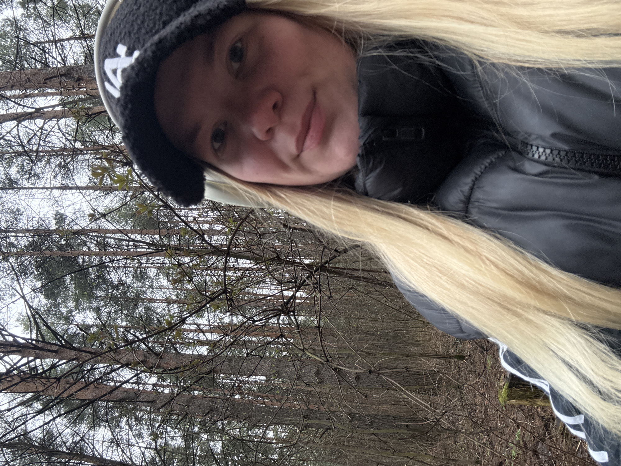 A selfie of a young woman with long, blonde hair, wearing a black beanie with white letters, a black padded jacket with white stripes on the sleeves. The background consists of a forest with bare trees. The location is likely near Chaikovsky, Russia based on the query.