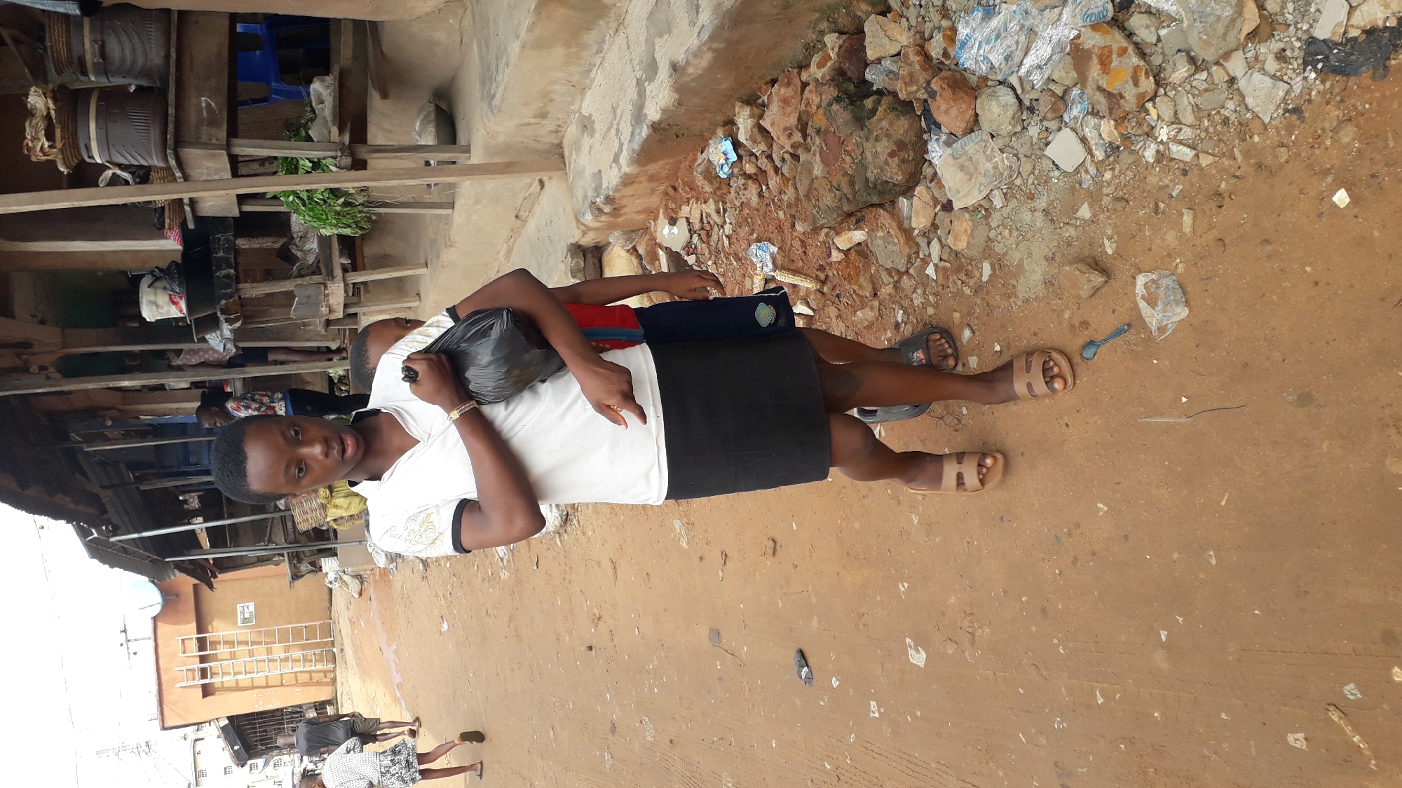 The image depicts a street scene in Nkpor, Nigeria, an area known for its bustling markets. In the foreground, a young person wearing a white t-shirt, a black skirt, and light brown sandals is walking towards the viewer, carrying a black plastic bag. To the right, a second, younger individual is partially visible, wearing a blue and red shirt and dark shorts. Behind them, a market stall with wooden shelves displays various items, including baskets and what appear to be food products. The ground is a mixture of dirt and rubble, with scattered stones and some plastic debris. In the background, a multi-story building with a ladder attached to its side is visible, suggesting a densely populated urban environment. The overall atmosphere suggests a typical day in a Nigerian market town.