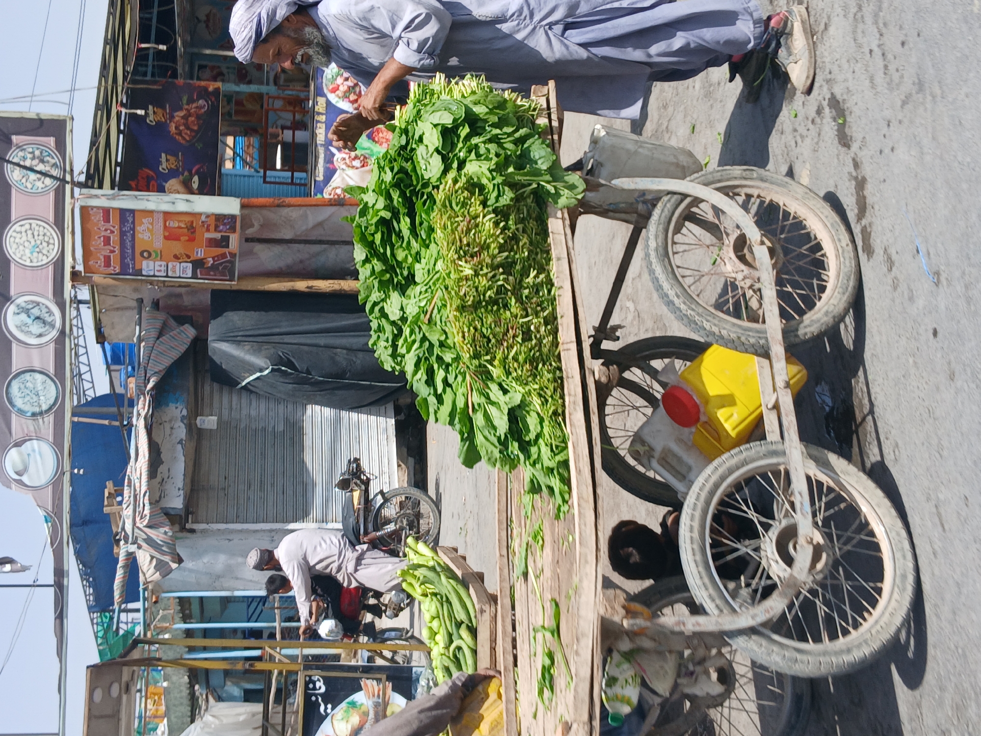 The image depicts a street scene in Quetta City Tehsil, Pakistan. An elderly man wearing a light-colored shalwar kameez and turban is arranging a large pile of green leafy vegetables, likely spinach or a similar local variety, on a wooden pushcart. The cart has bicycle-style wheels and appears to be handmade. Next to the spinach is a smaller pile of long, green cucumbers or gourds.
Behind the cart and the vendor are storefronts with signs in Urdu. One sign features images of food, possibly advertisements for a local restaurant. Another man, also wearing a shalwar kameez, is attending to a motorcycle parked near one of the shops. The area seems to be a bustling market street.