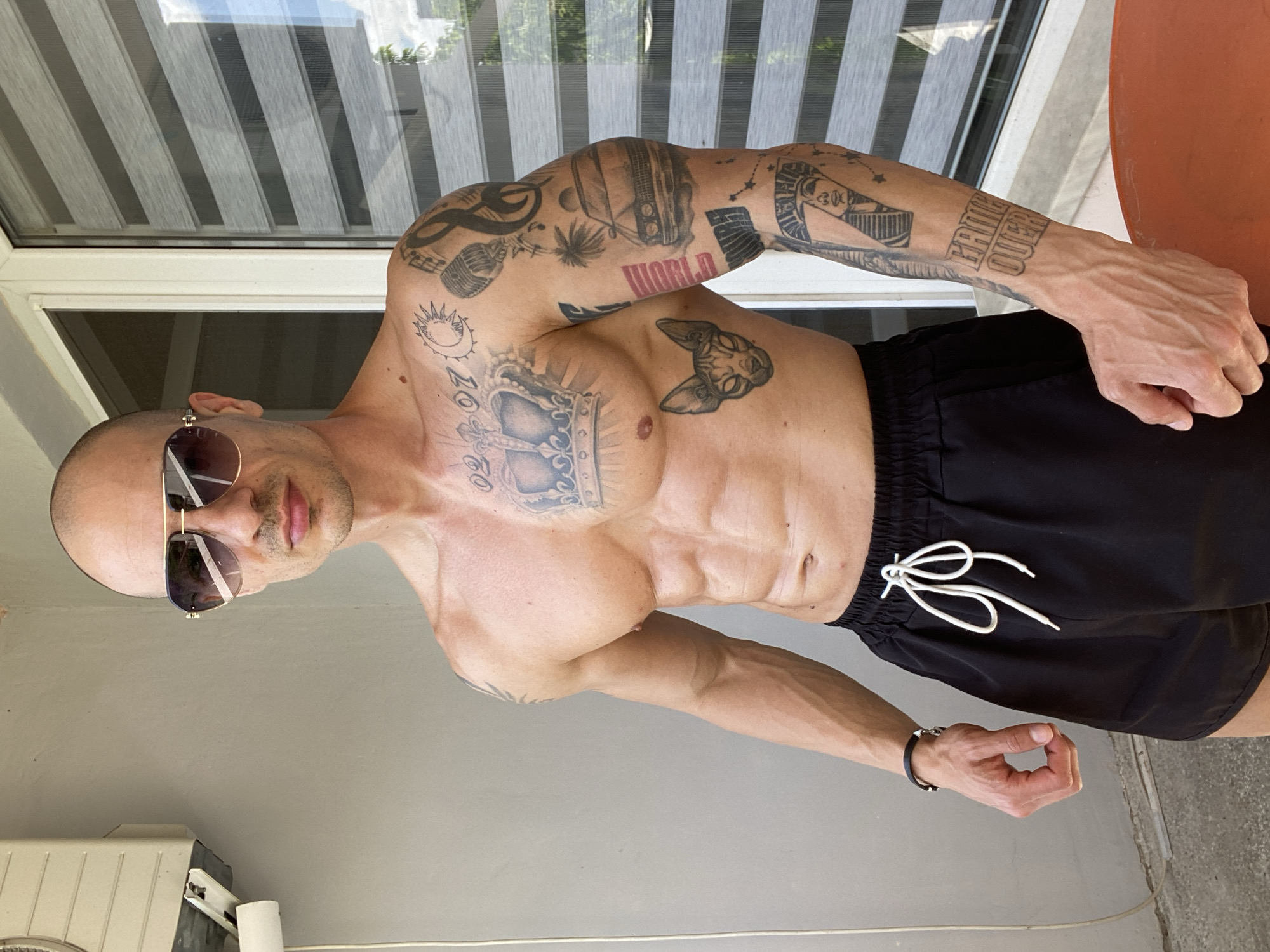 A shirtless, bald man with numerous tattoos is the central subject. He wears sunglasses and black shorts with a white drawstring. He is indoors, near a window with blinds. The location is Pleven, Bulgaria.